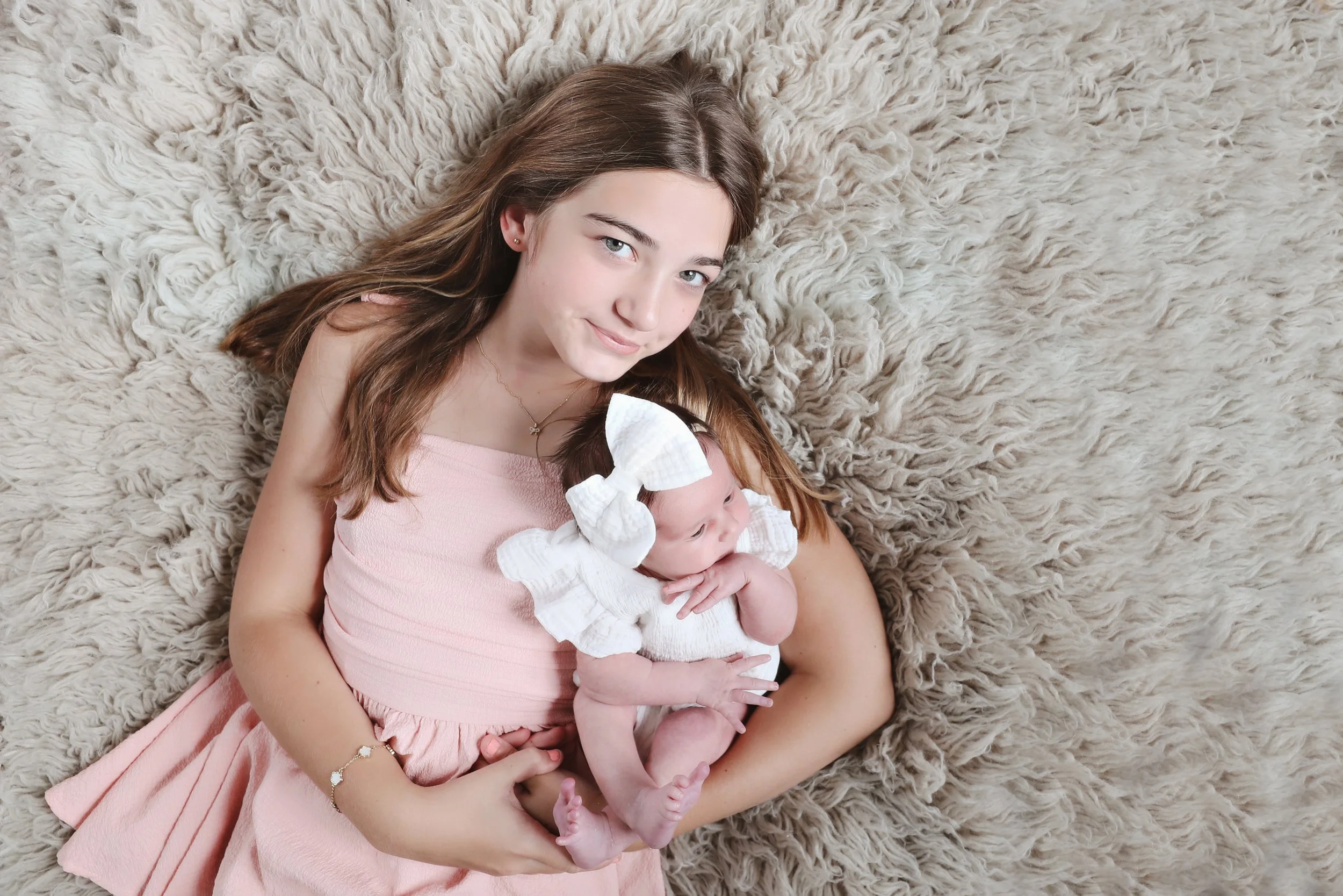 A young girl with long brown hair lying on a fluffy beige rug, holding a small baby dressed in white with a large white bow on their head.