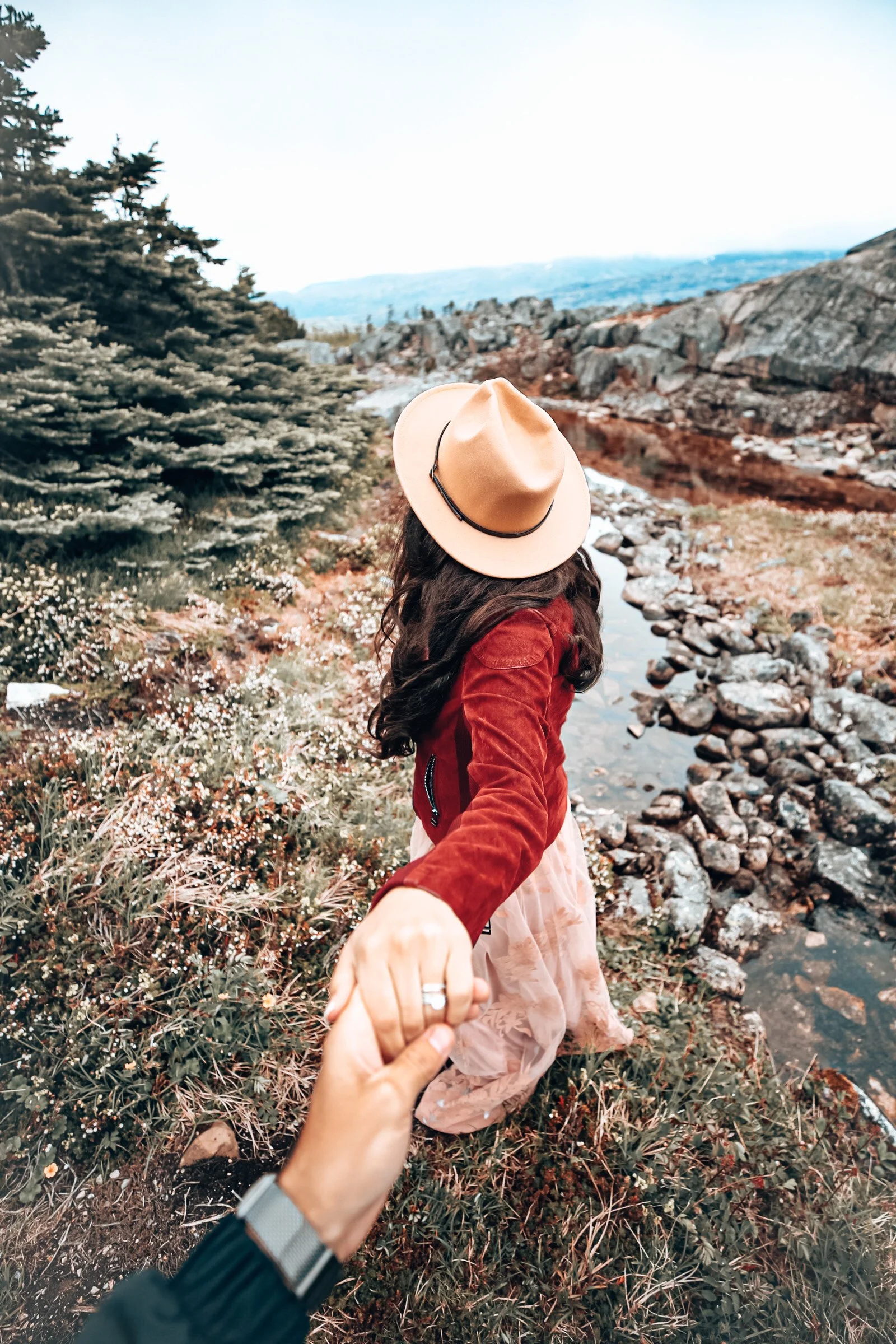 A woman in a beige hat and red jacket holding hands with someone while standing near a rocky creek in a forested area.