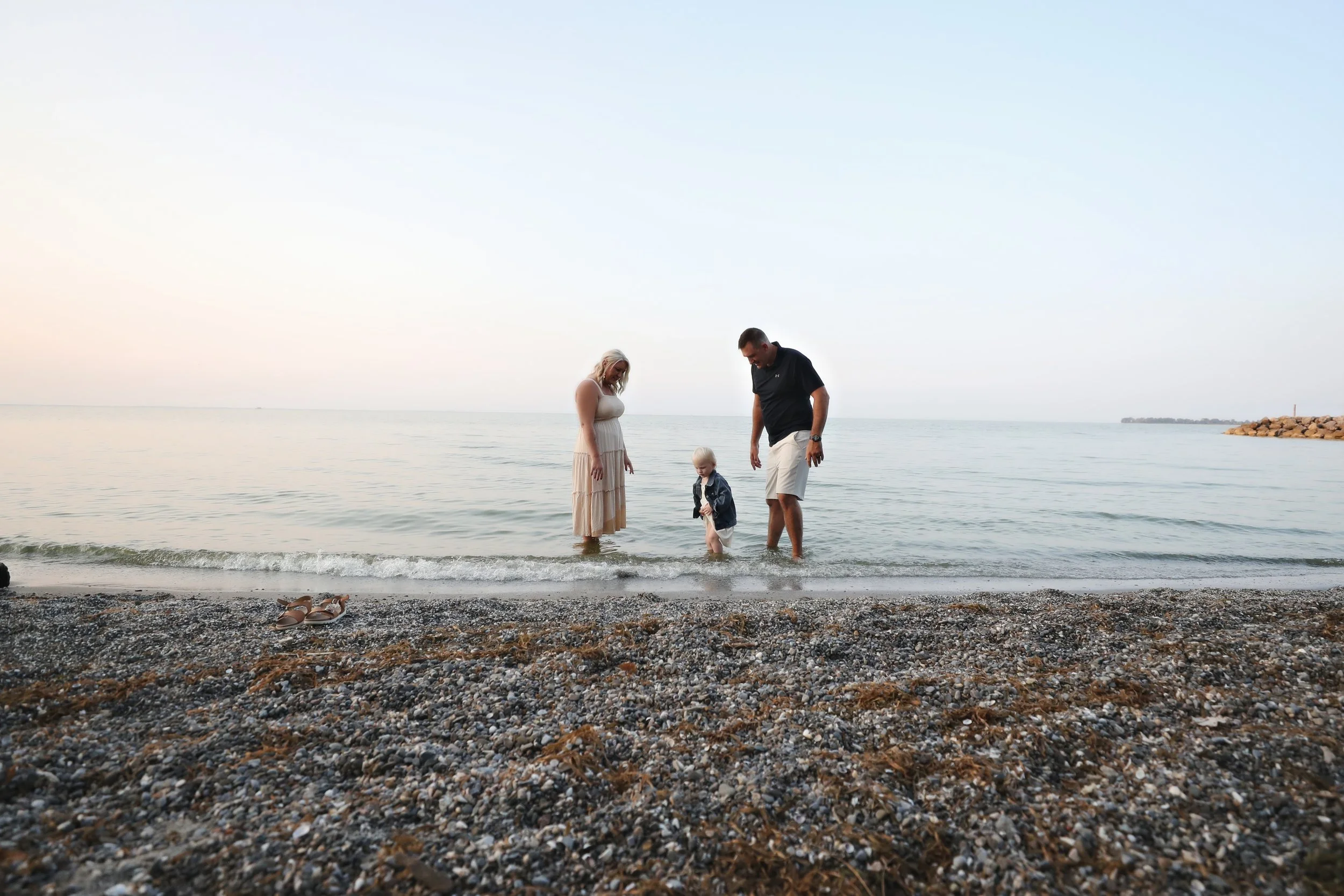 A family at the beach, two adults and a child, standing in shallow water with the shoreline and a clear sky in the background.