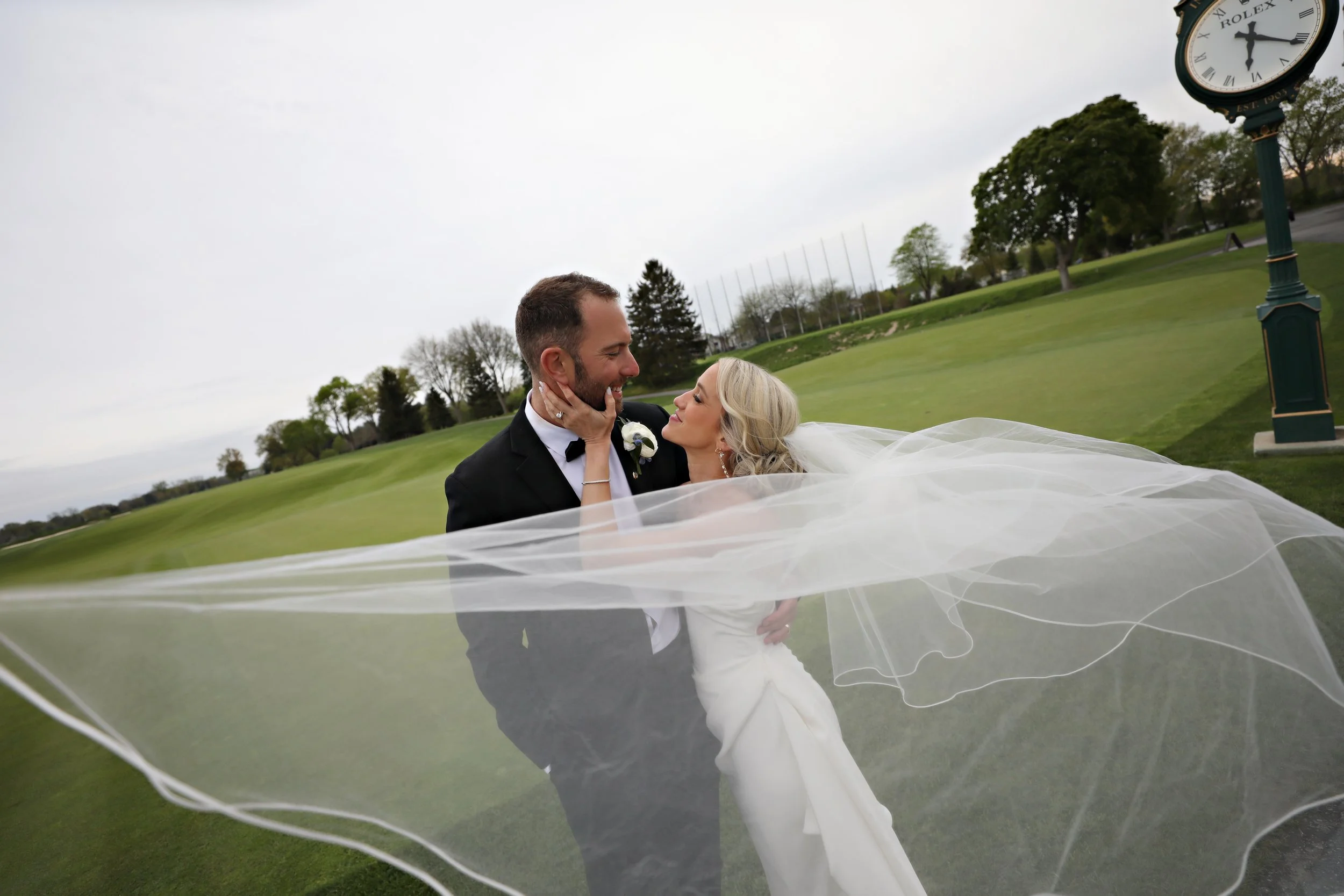 A bride and groom on a golf course, embracing and gazing at each other, with the bride's veil flowing around them and a clock tower in the background.