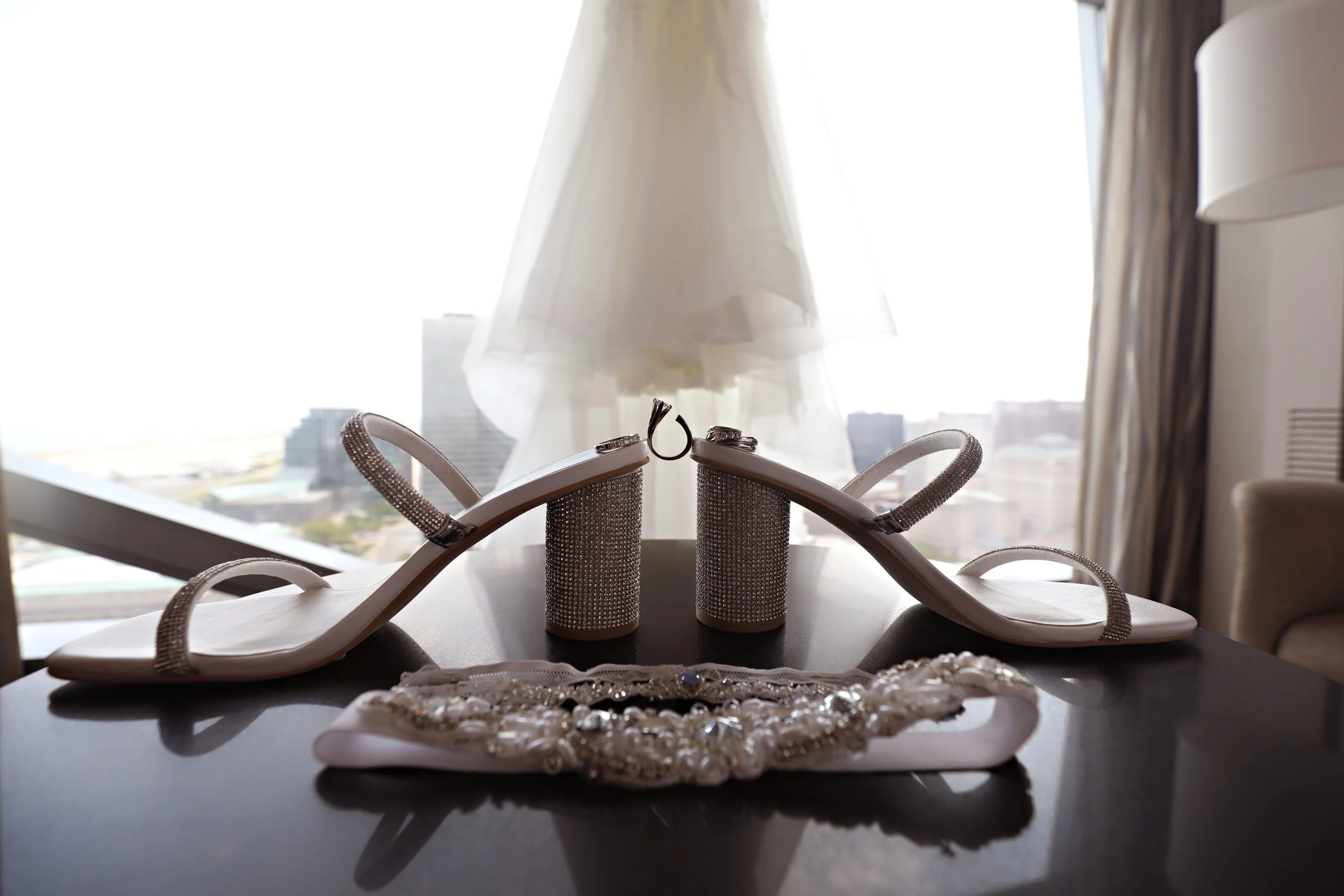 A pair of white high-heeled sandals with rhinestone straps, a wedding dress on a hanger, a pearl and rhinestone necklace, and wedding rings on a dark table with a city view in the background.