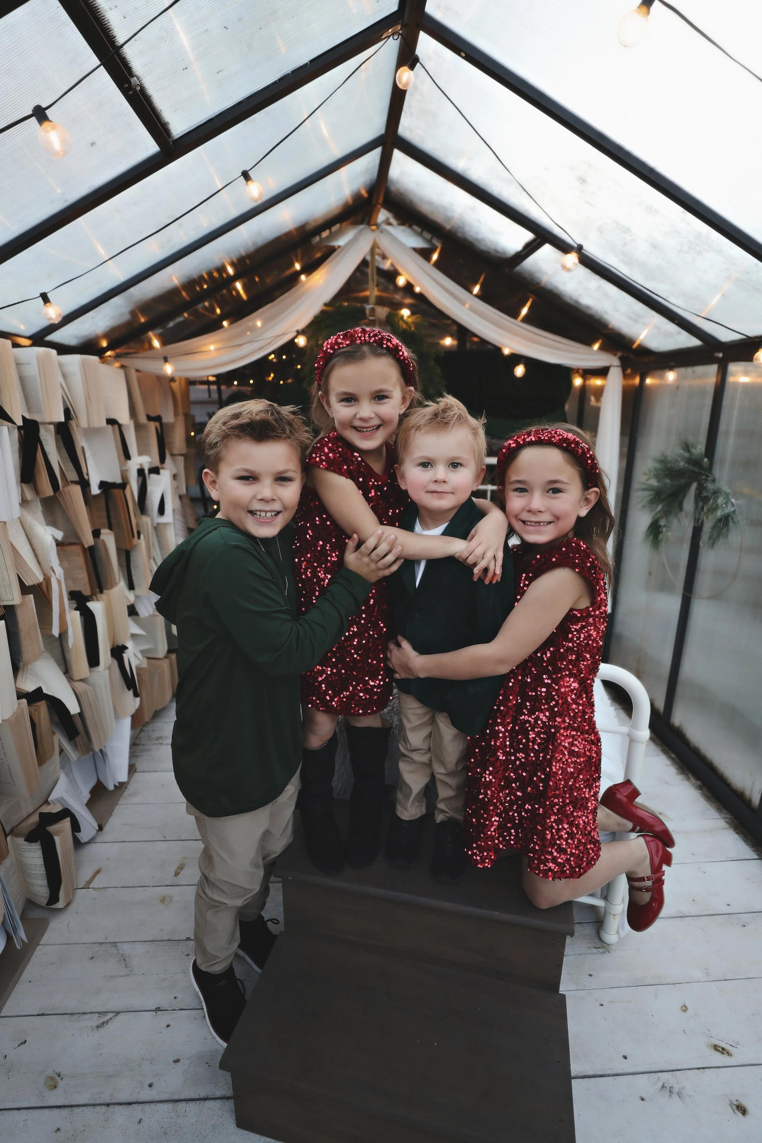 Group of five children, dressed in festive clothing, happily posing inside a decorated greenhouse or conservatory with string lights and greenery.