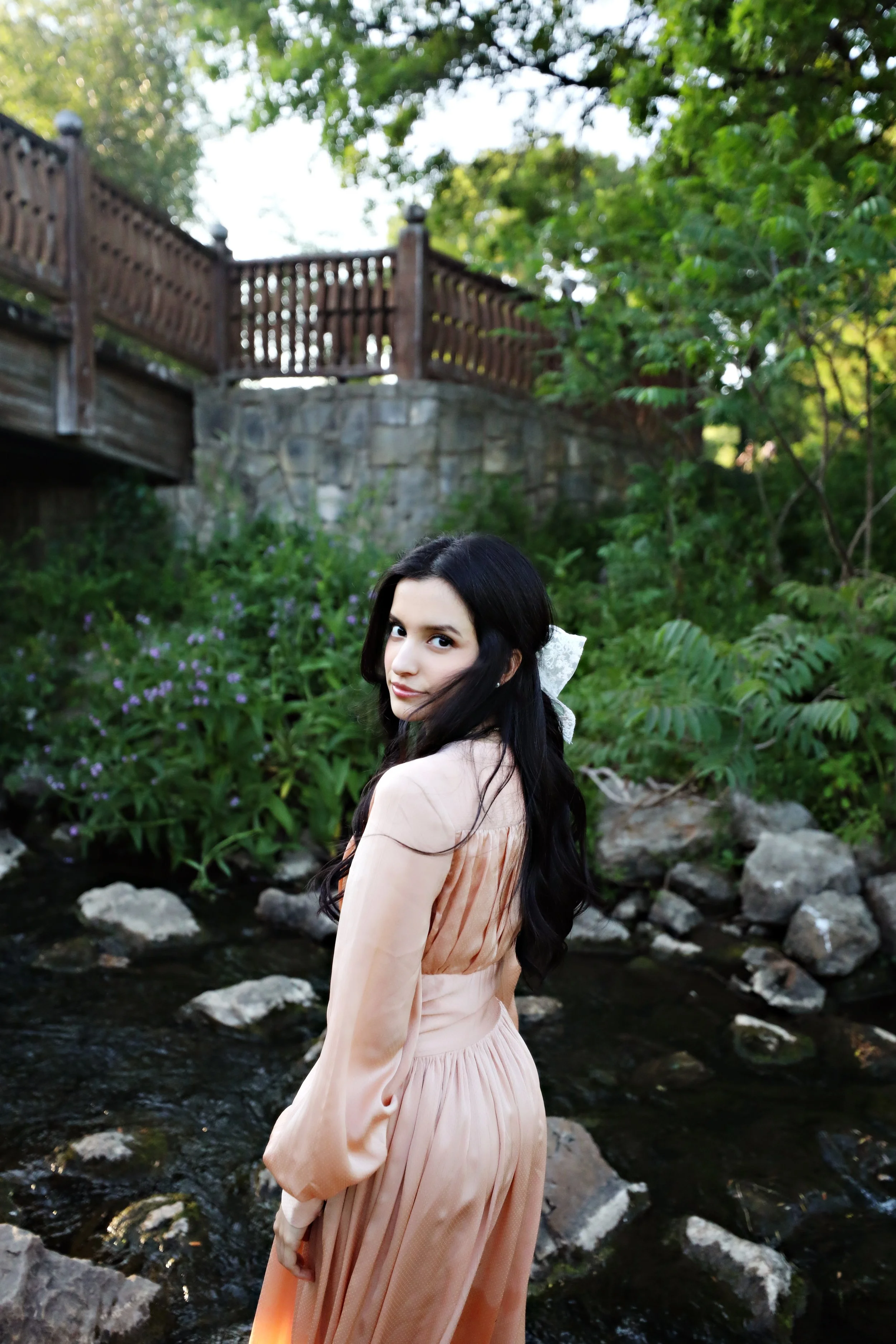 Young woman with long dark hair wearing a peach-colored dress standing in a garden with a stream, rocks, green plants, and a wooden bridge in the background.