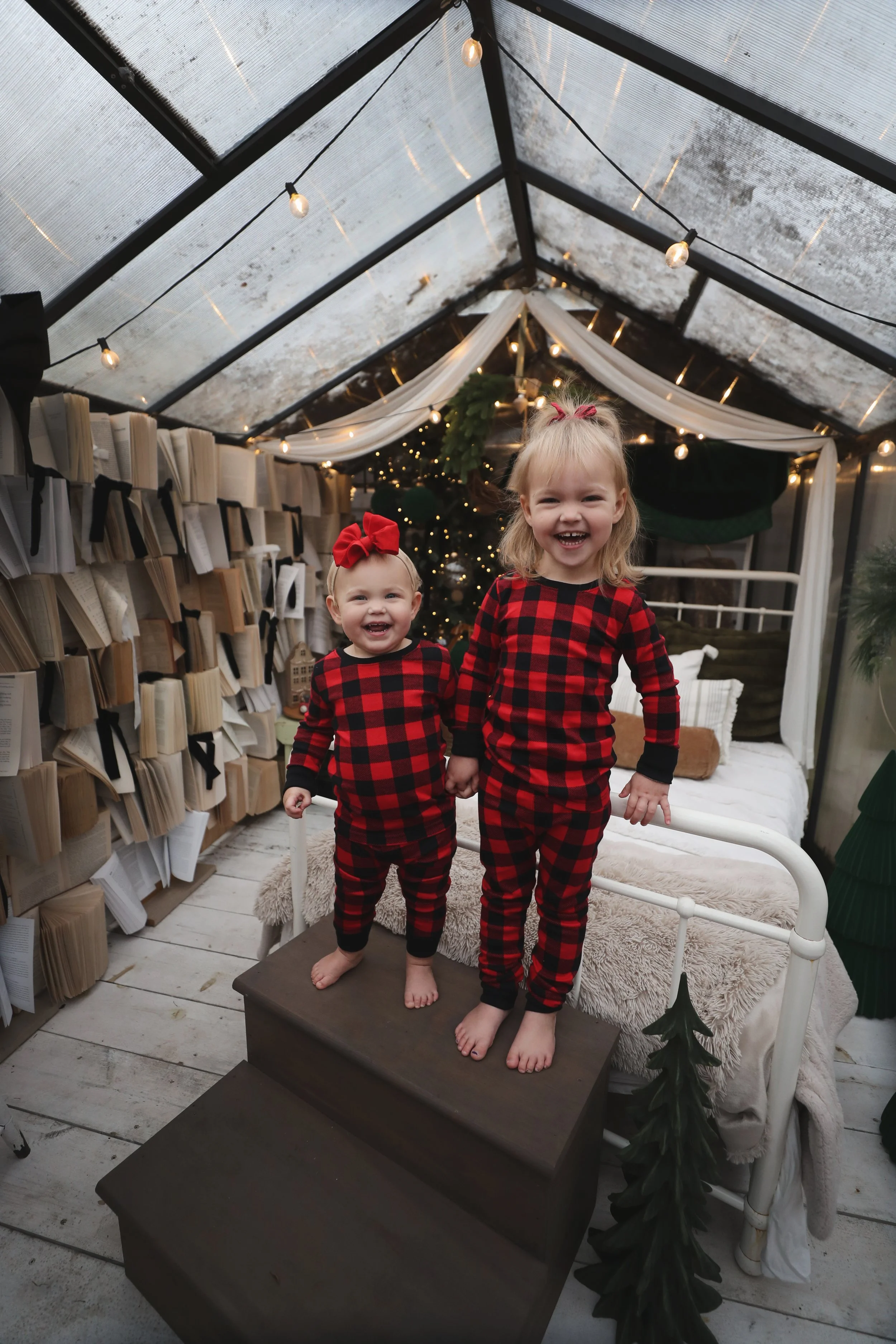 Two young girls in matching red and black plaid pajamas standing on a small staircase inside a decorated glass-enclosed room, smiling and holding hands, with a bed, Christmas tree, and Christmas lights in the background.