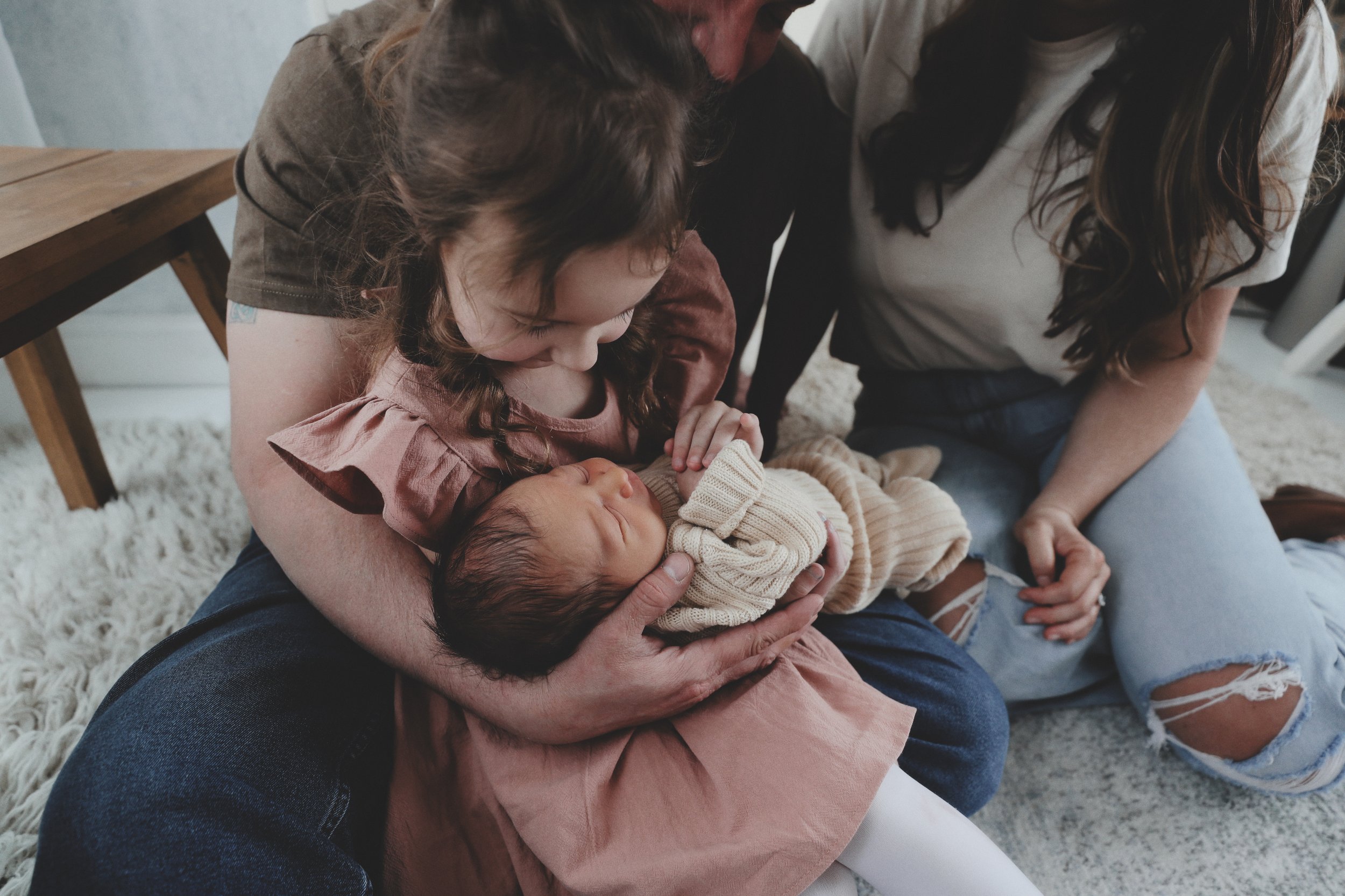 A woman holding a newborn baby while a girl looks over and touches the baby's forehead in a cozy home setting.
