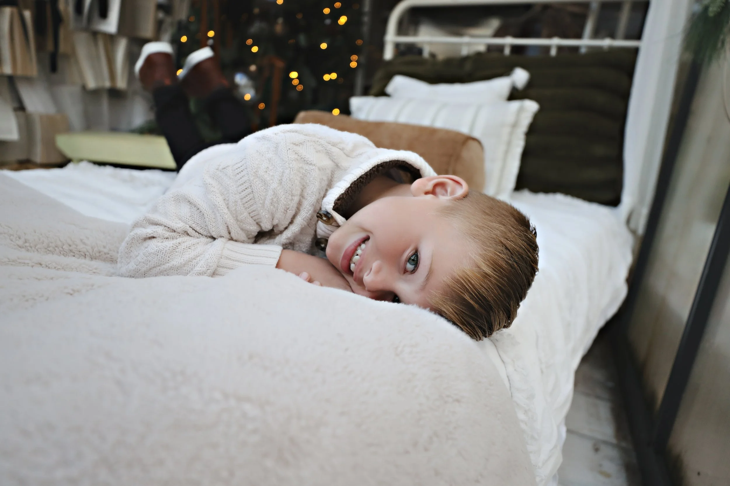 A young boy with brown hair and blue eyes lying on a bed, smiling and looking at the camera. He is wearing a white knit sweater and is in a cozy room with bookshelves, pillows, and a background of blurred warm yellow lights.