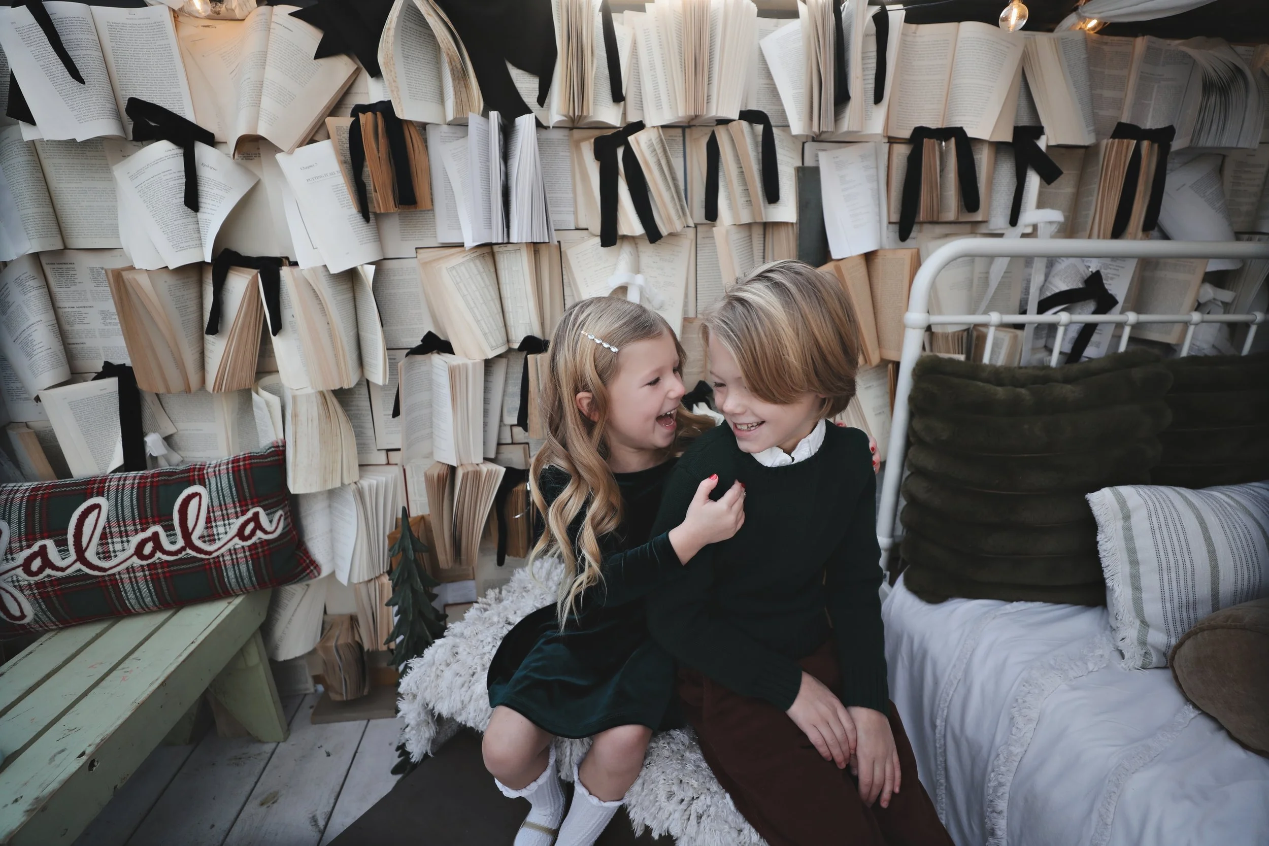 Two children, a girl and a boy, sitting close together on a fluffy blanket, smiling and laughing, in a cozy room with a wall decorated with open books tied with black ribbons, a Christmas pillow with 