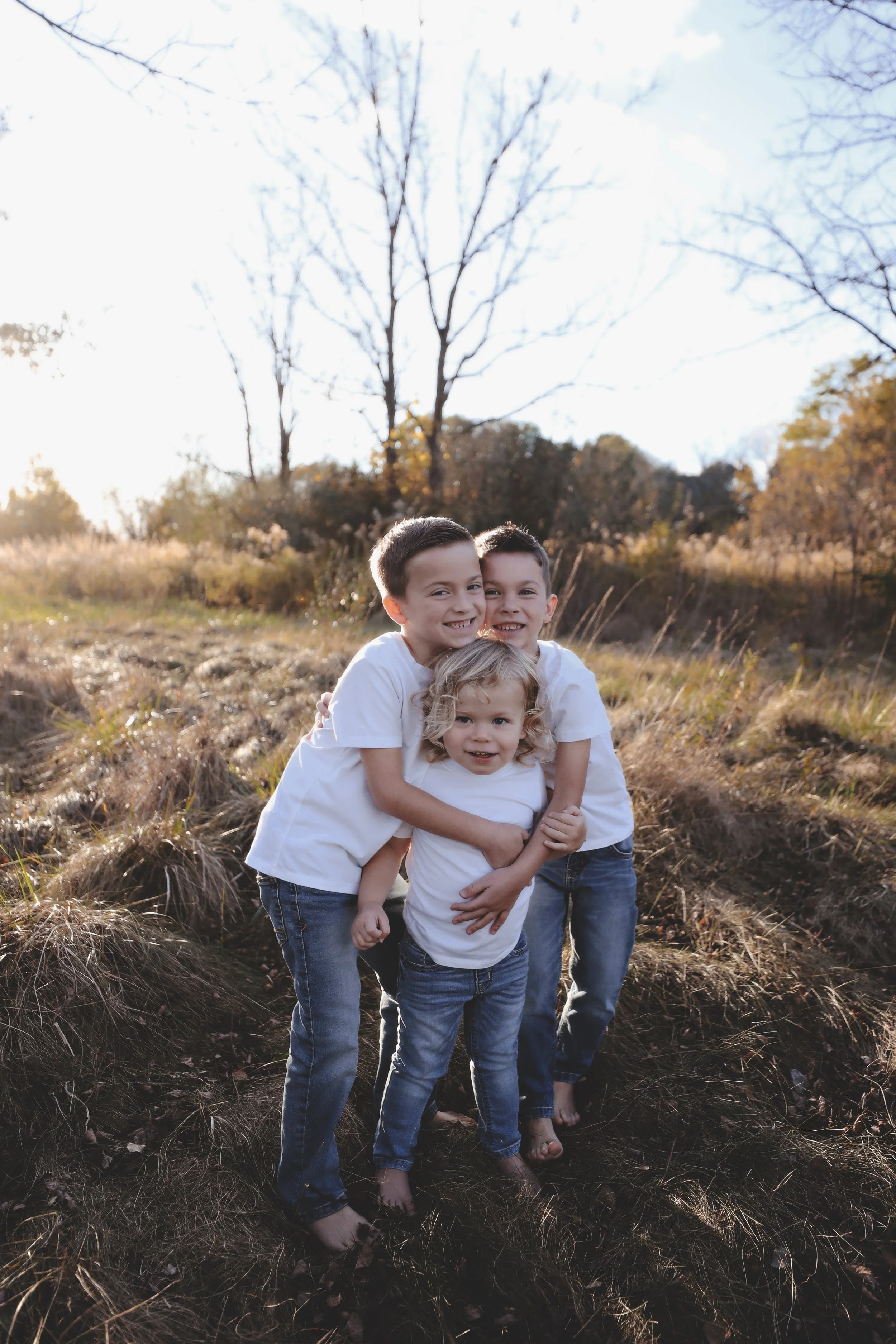 Three children, two boys and a girl, hugging each other outdoors in a field with autumn trees in the background, wearing white t-shirts and jeans.