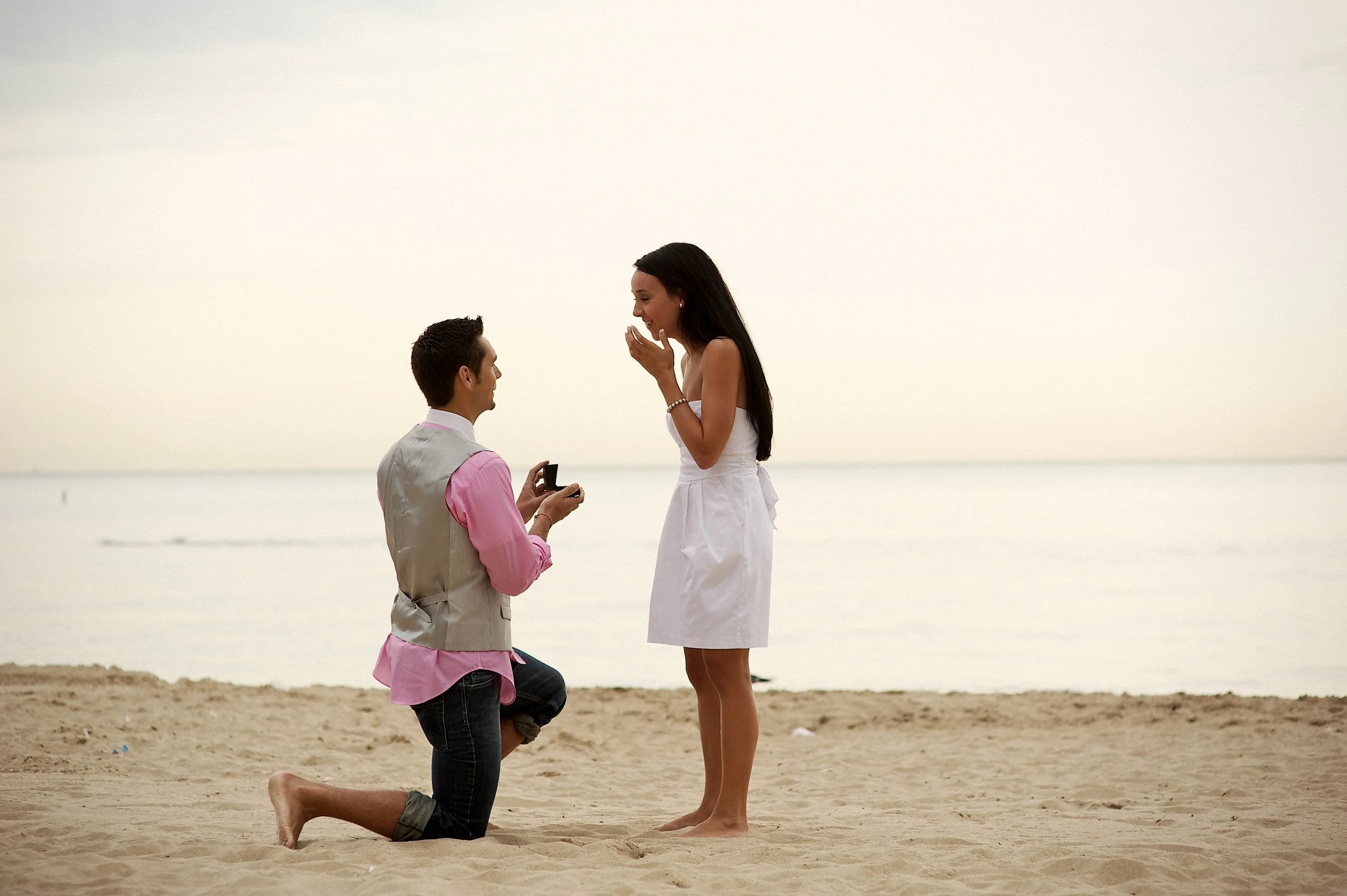 A man proposing marriage to a woman on the beach at sunset, with the man kneeling and the woman smiling and covering her mouth.