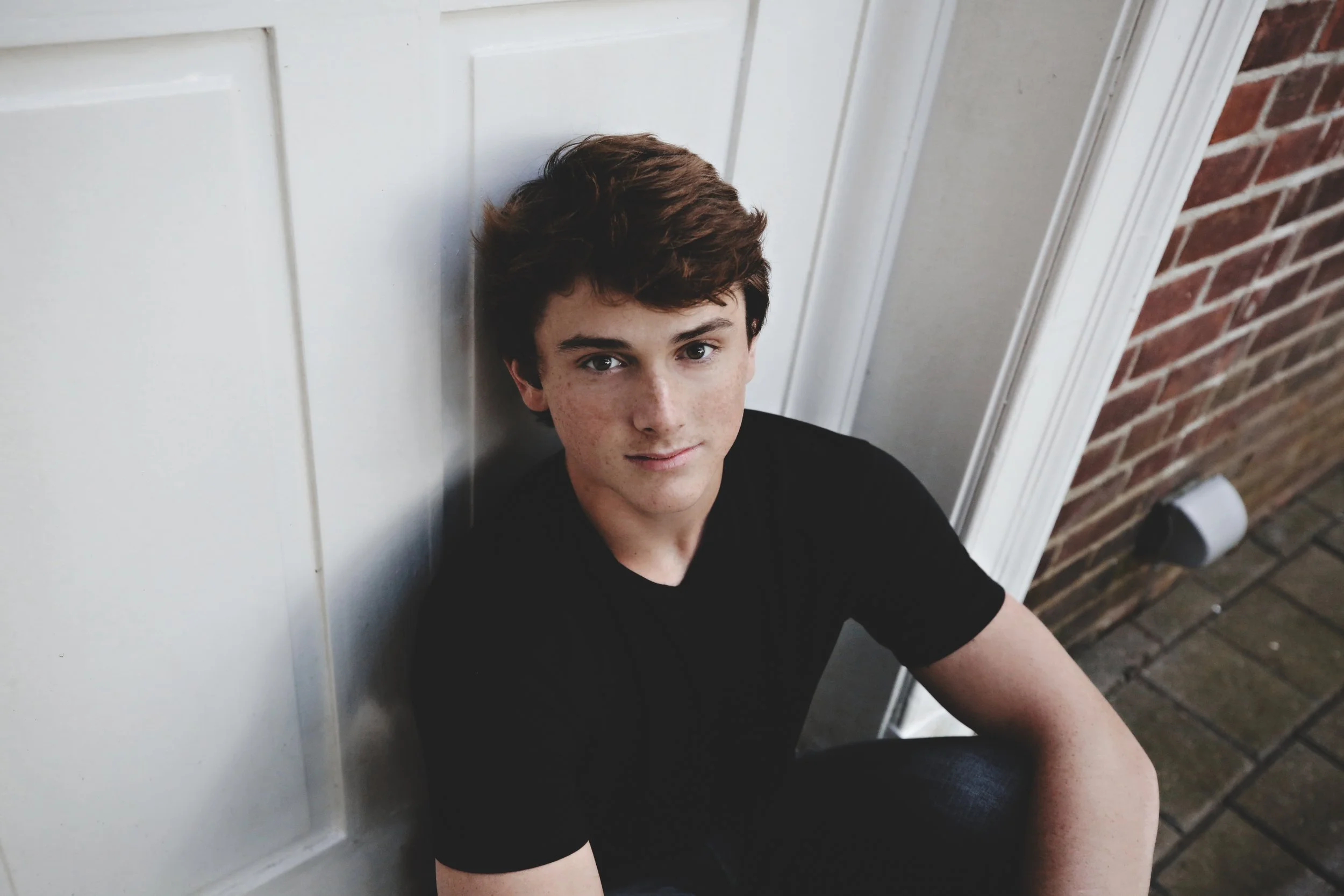 A young boy with brown hair, wearing a black shirt, sitting against a white wall next to a brick wall outside.