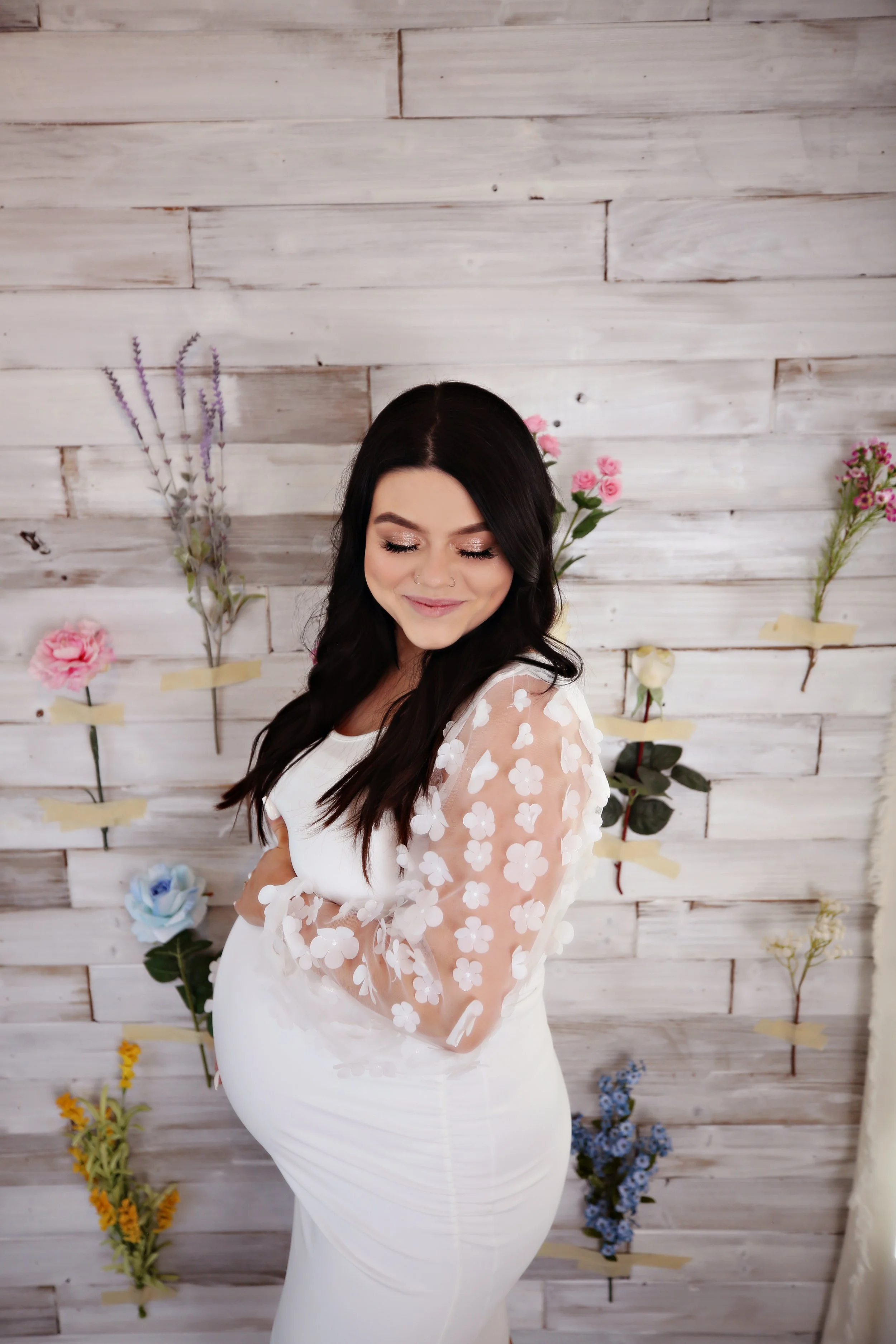 Pregnant woman in a white dress with floral sheer sleeves, standing against a rustic white wood wall decorated with various colorful artificial flowers.