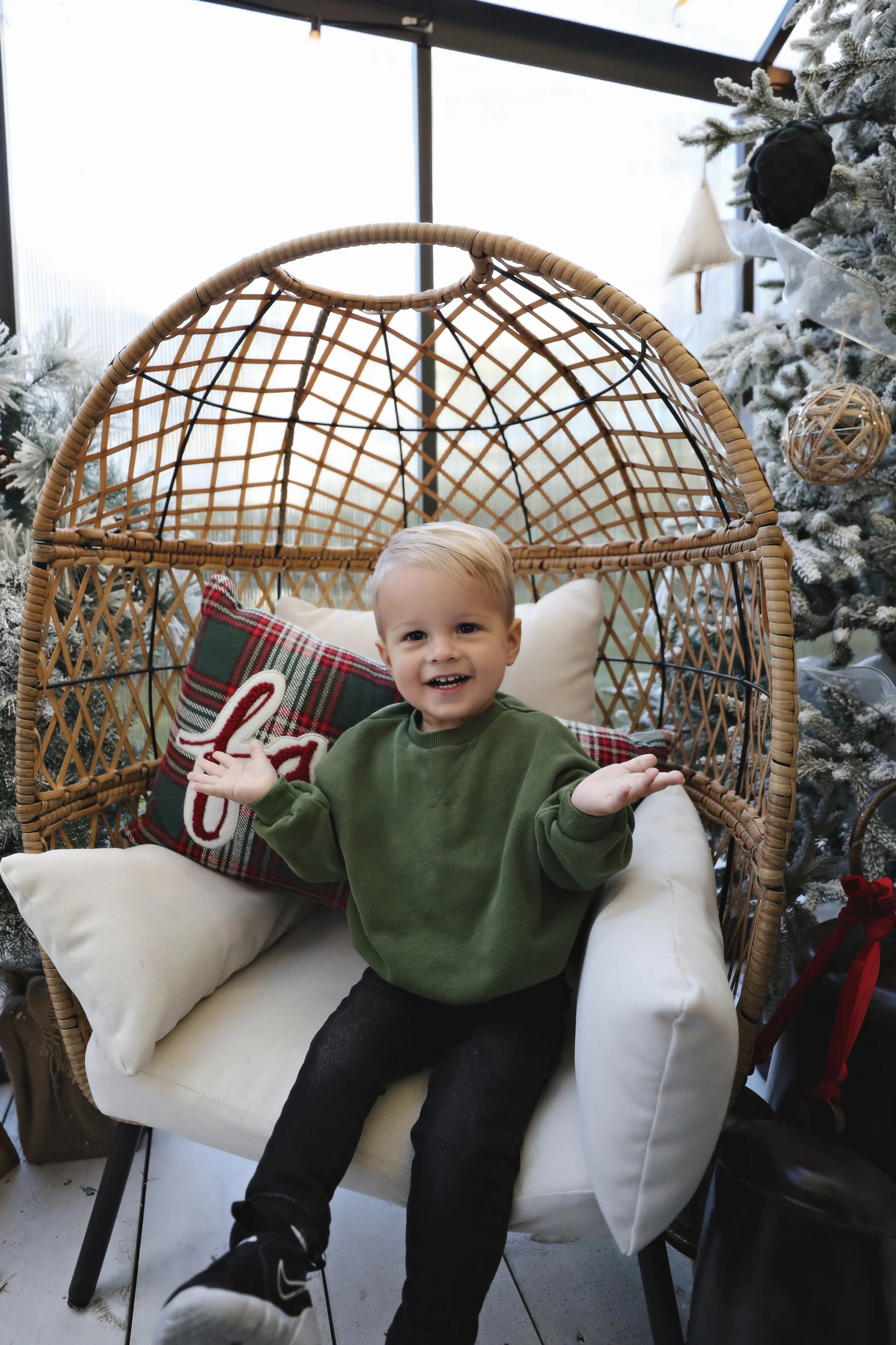 A young boy with blonde hair, wearing a green sweatshirt and black pants, sitting on a cushioned chair with a rattan back, near a decorated Christmas tree, smiling and gesturing with his hands.