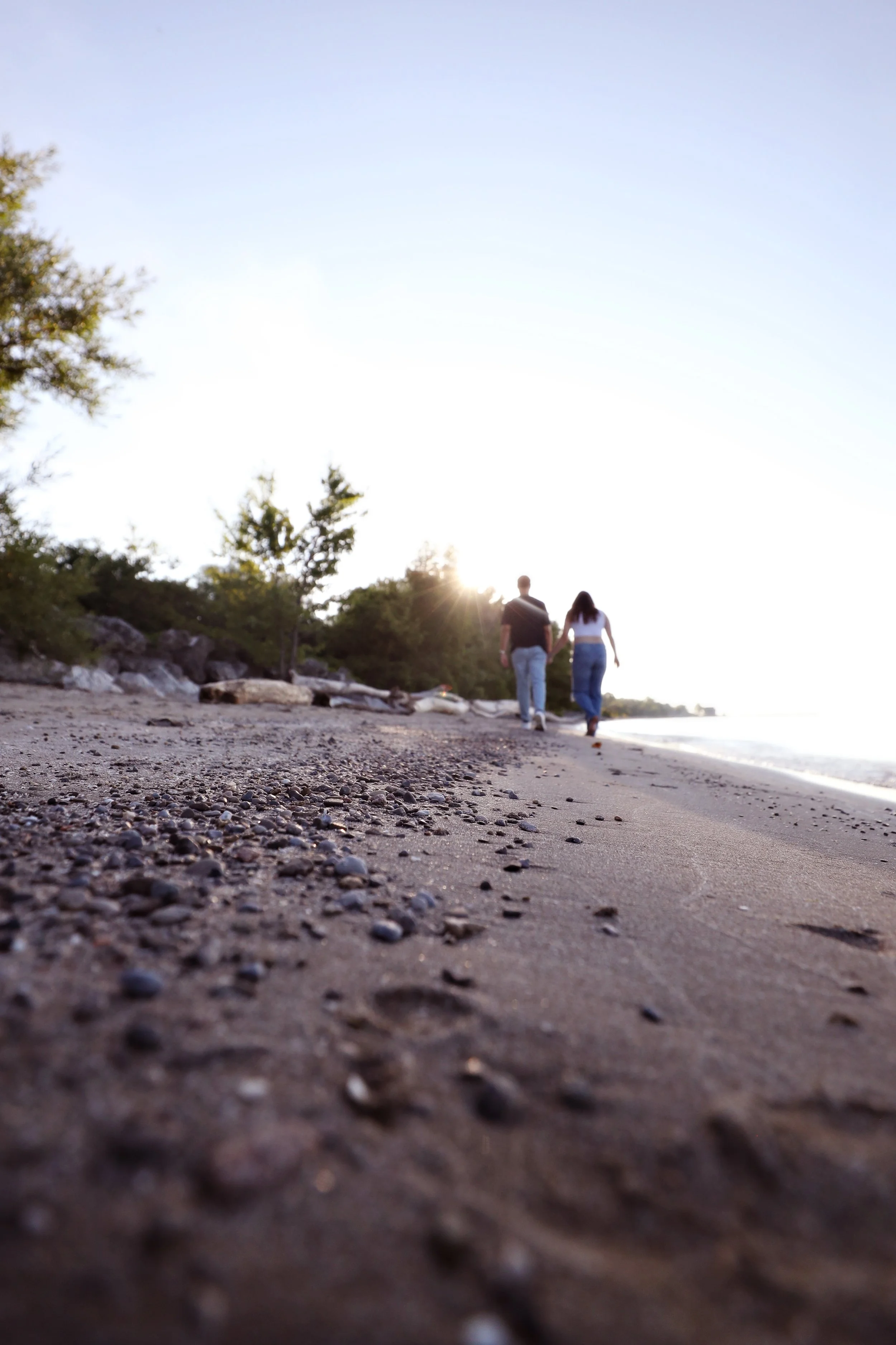 A couple walking hand in hand along a sandy beach at sunset, with trees on one side and the ocean on the other.