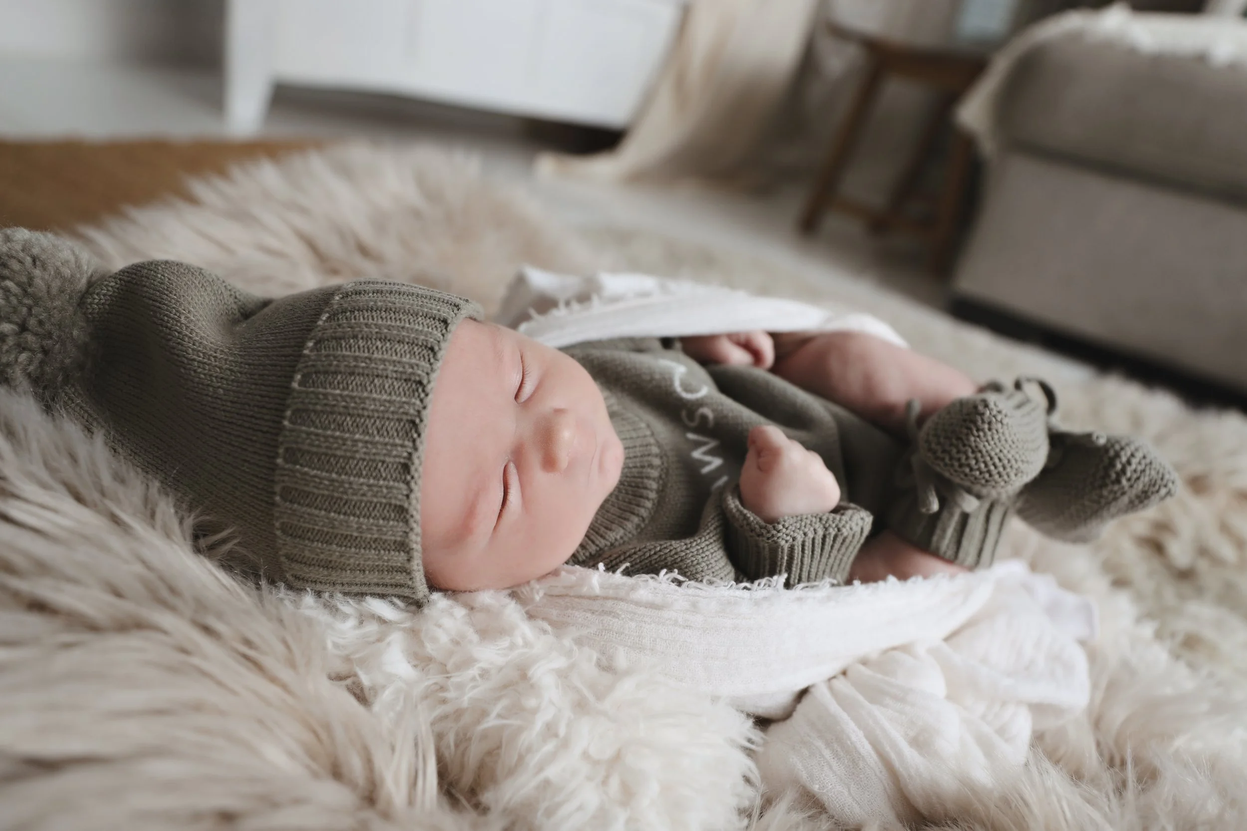 A sleeping baby wearing a gray knit beanie, matching mittens, and a cozy outfit, lying on a soft, fluffy rug.