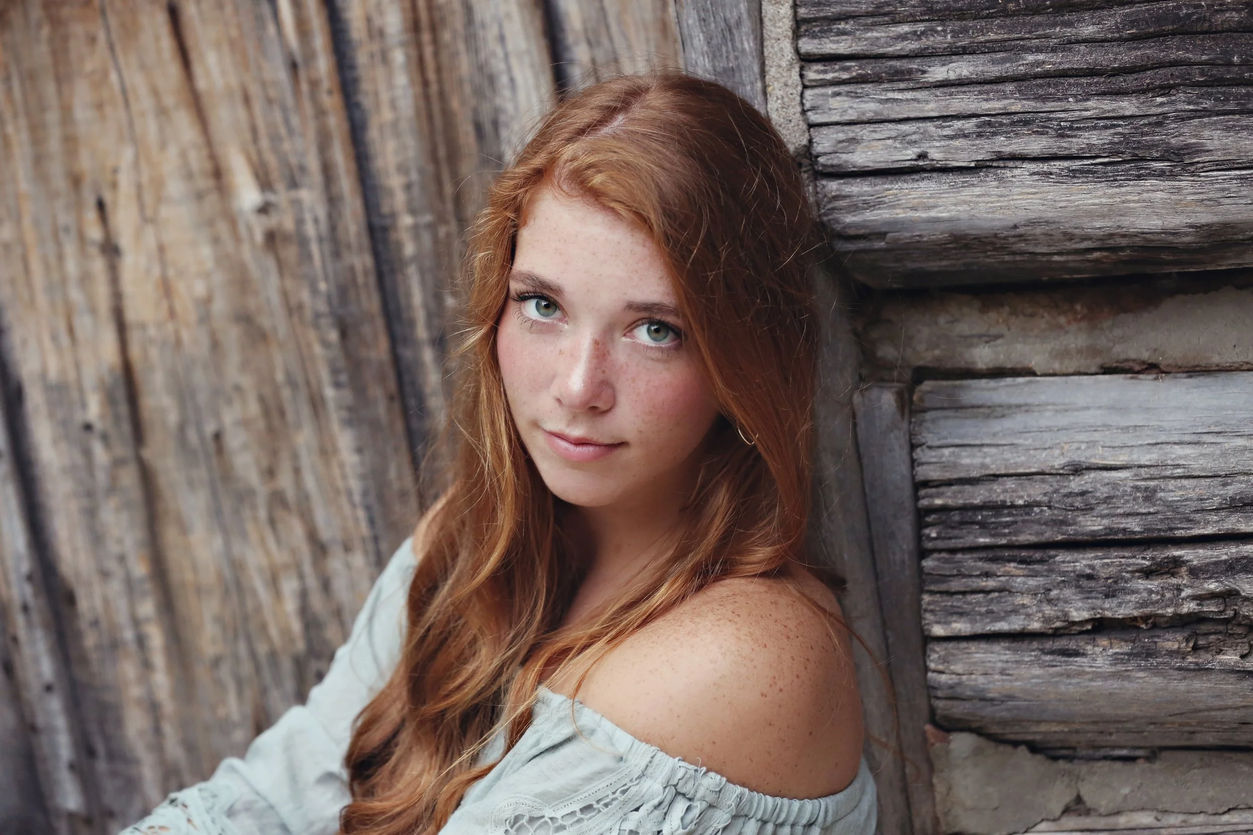 A young woman with red hair and blue eyes leaning against a rustic wooden wall, looking at the camera.