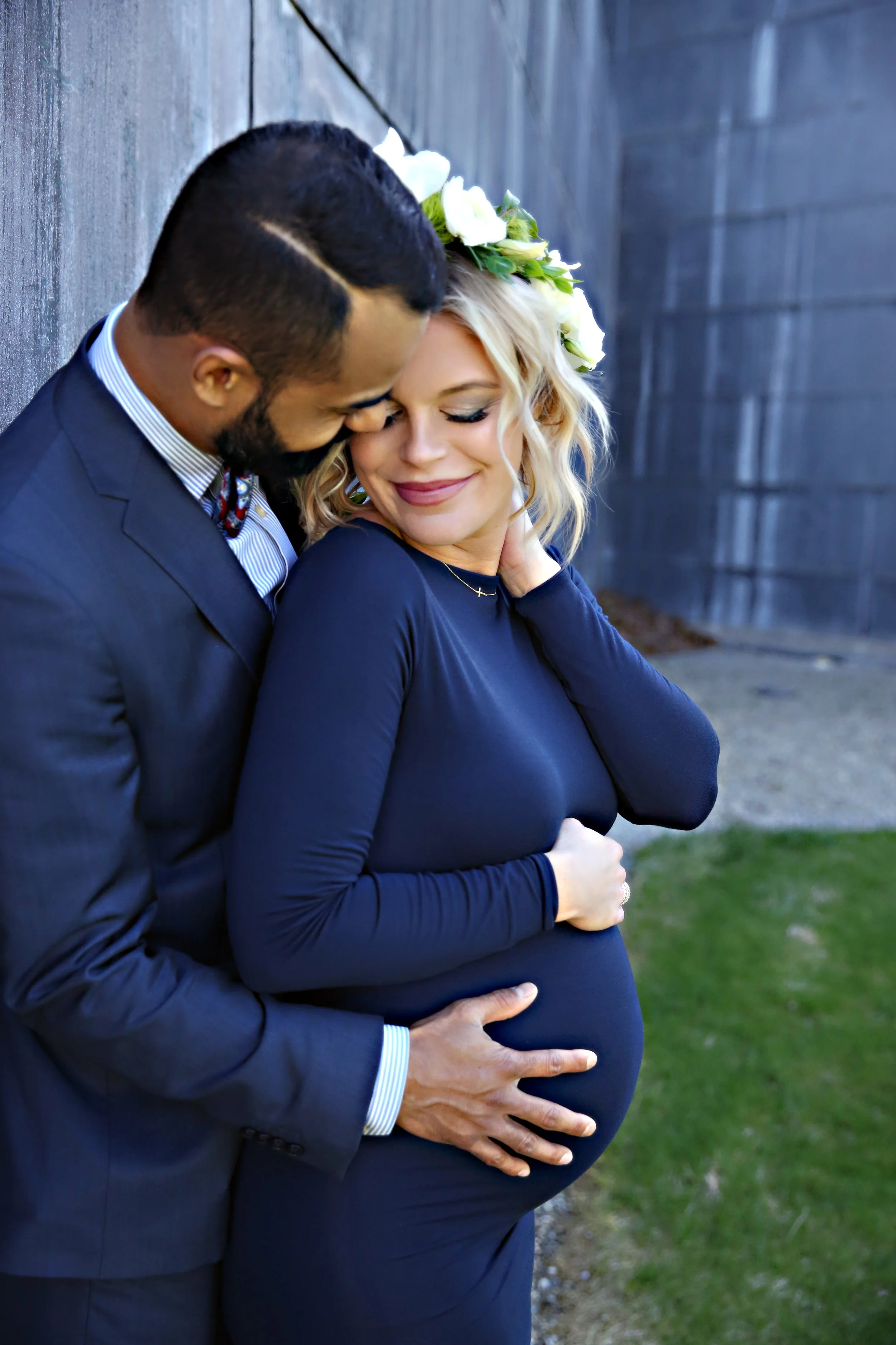 A couple stands close together, with the man touching the pregnant woman's belly. The woman has blonde, curly hair and a white floral crown, and is smiling with eyes closed. The man has short, dark hair and a beard, and is wearing a navy suit with a 