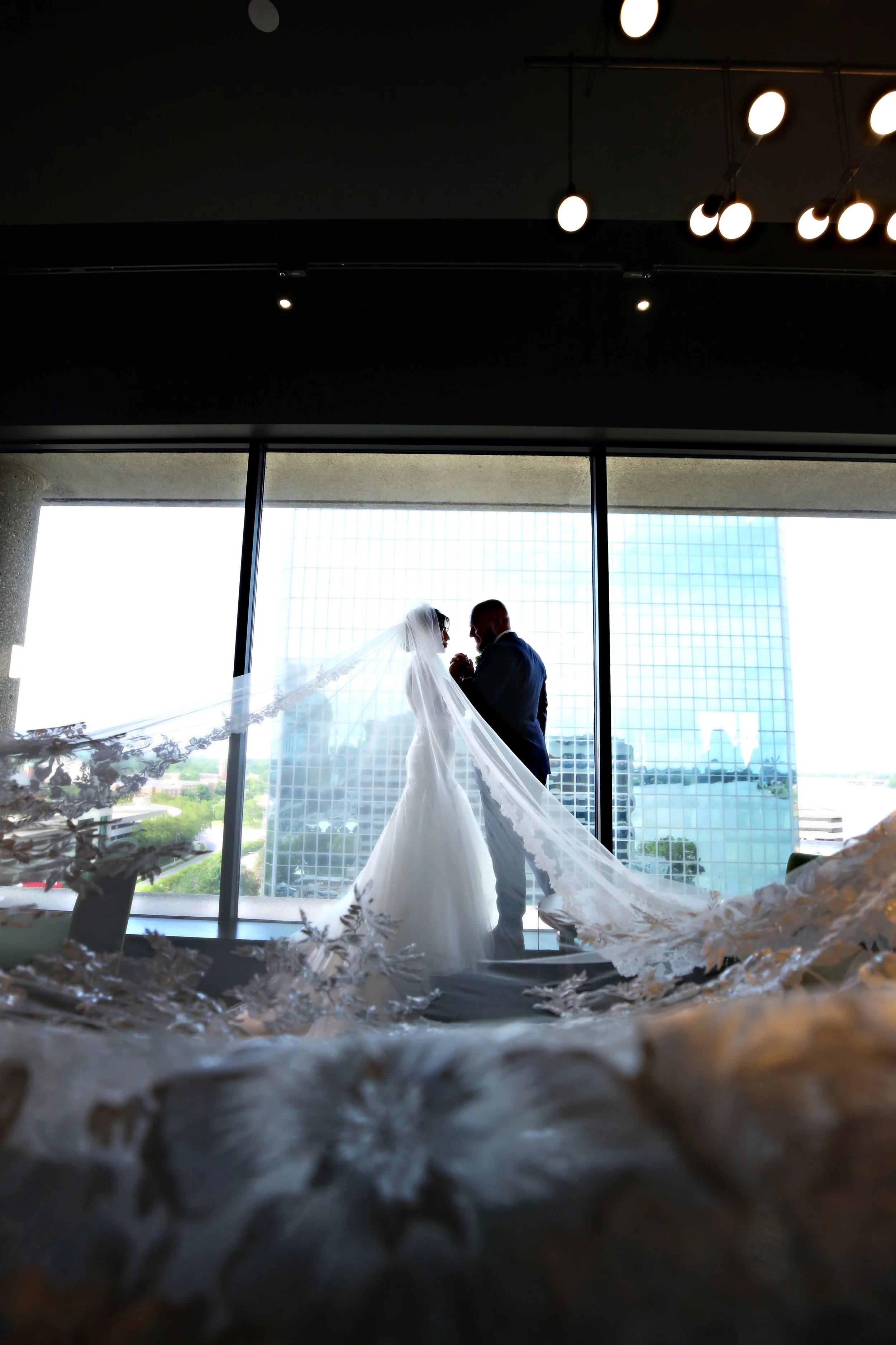 A bride and groom standing close together in front of a large window, sharing a moment during their wedding day.
