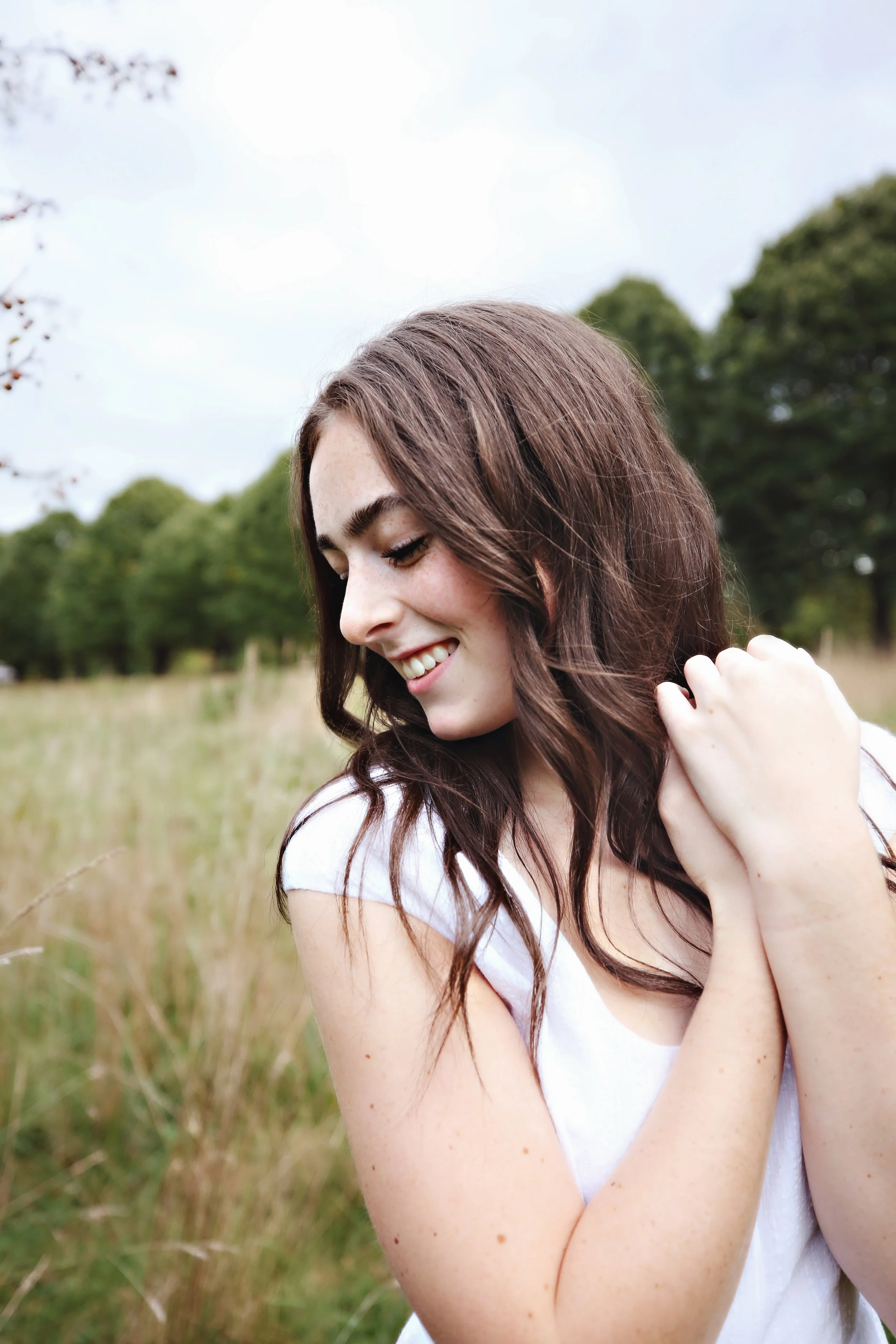 A young woman with long brown hair smiling with closed eyes, standing in a field of tall grass with trees in the background on a partly cloudy day.