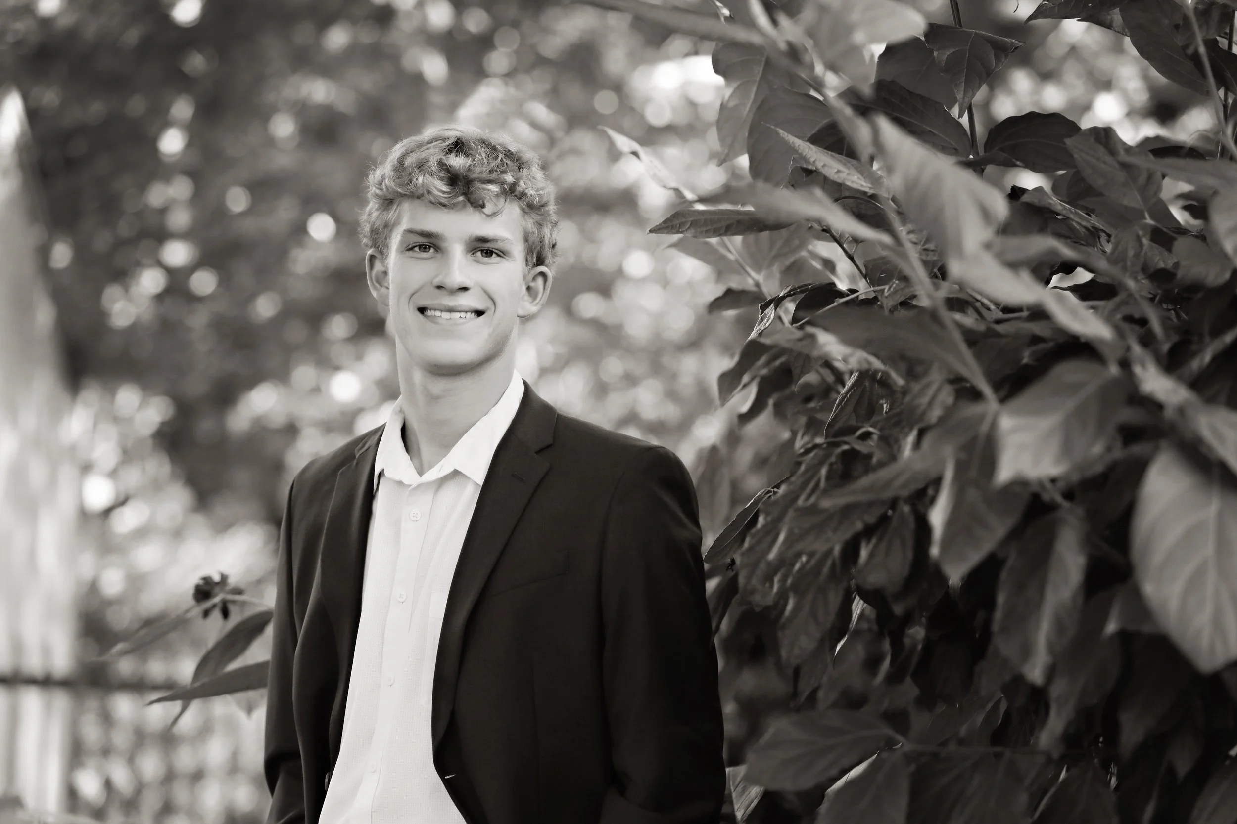 A young man in a suit and white shirt standing outdoors in front of trees and foliage, smiling at the camera, black and white photo.