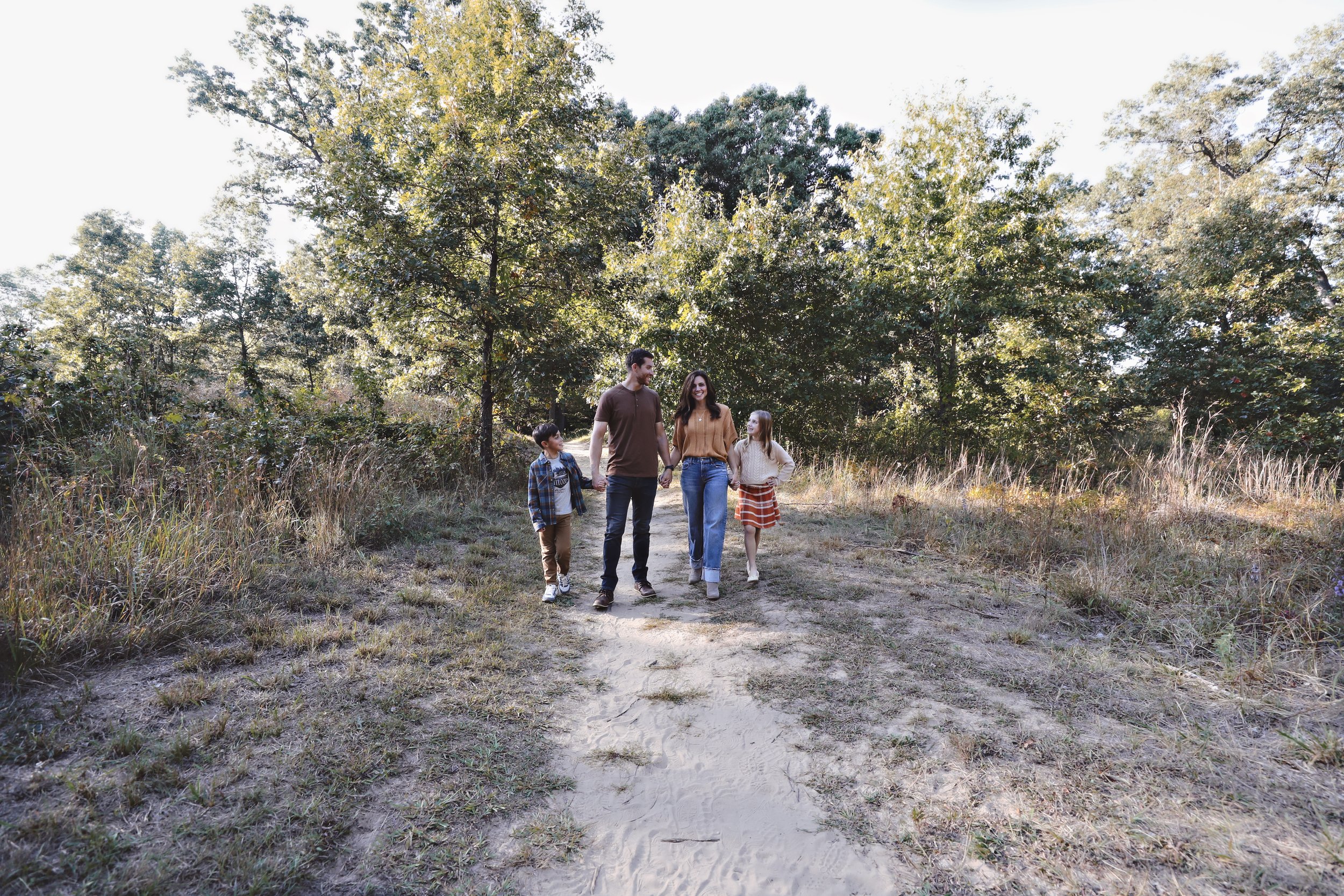 A family of four walking on a dirt trail through a wooded area on a sunny day.