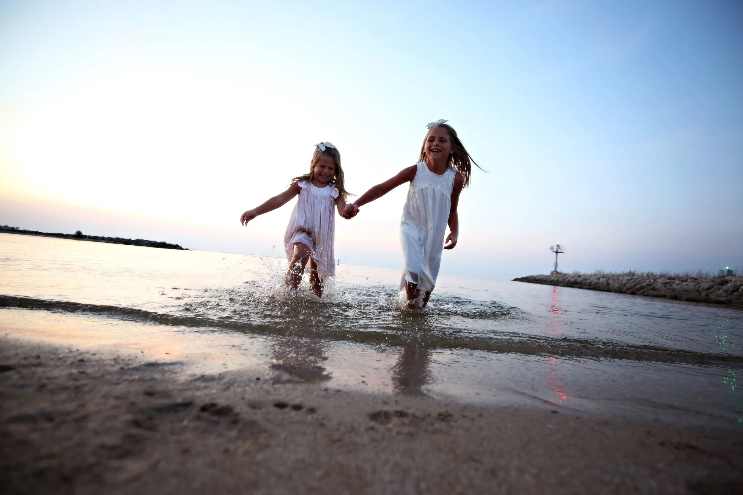 Two young girls in white dresses running and playing in the shallow water at the beach during sunset, holding hands and smiling.
