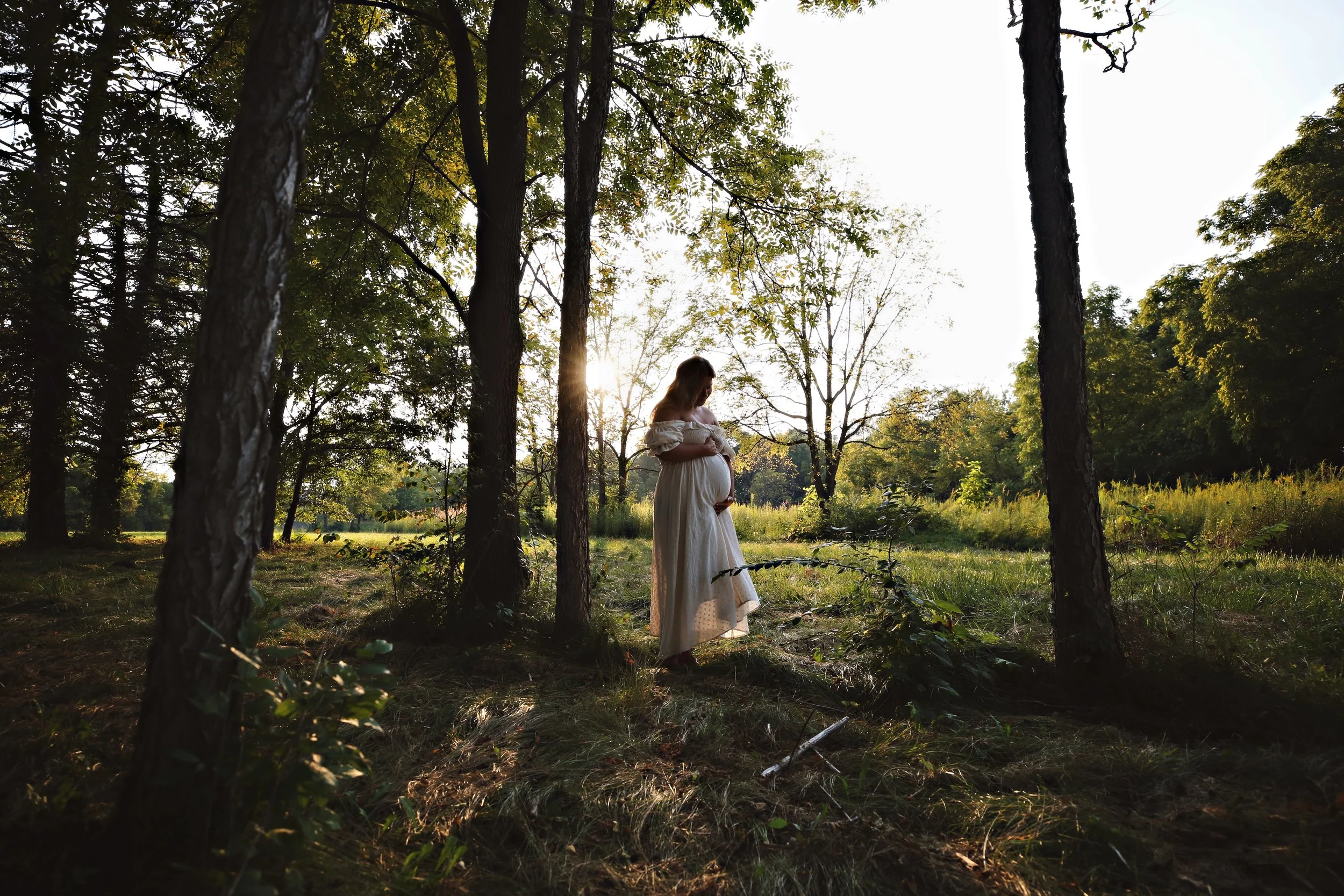 Pregnant woman in a flowing white dress standing in a wooded area at sunset, cradling her baby bump and looking down.