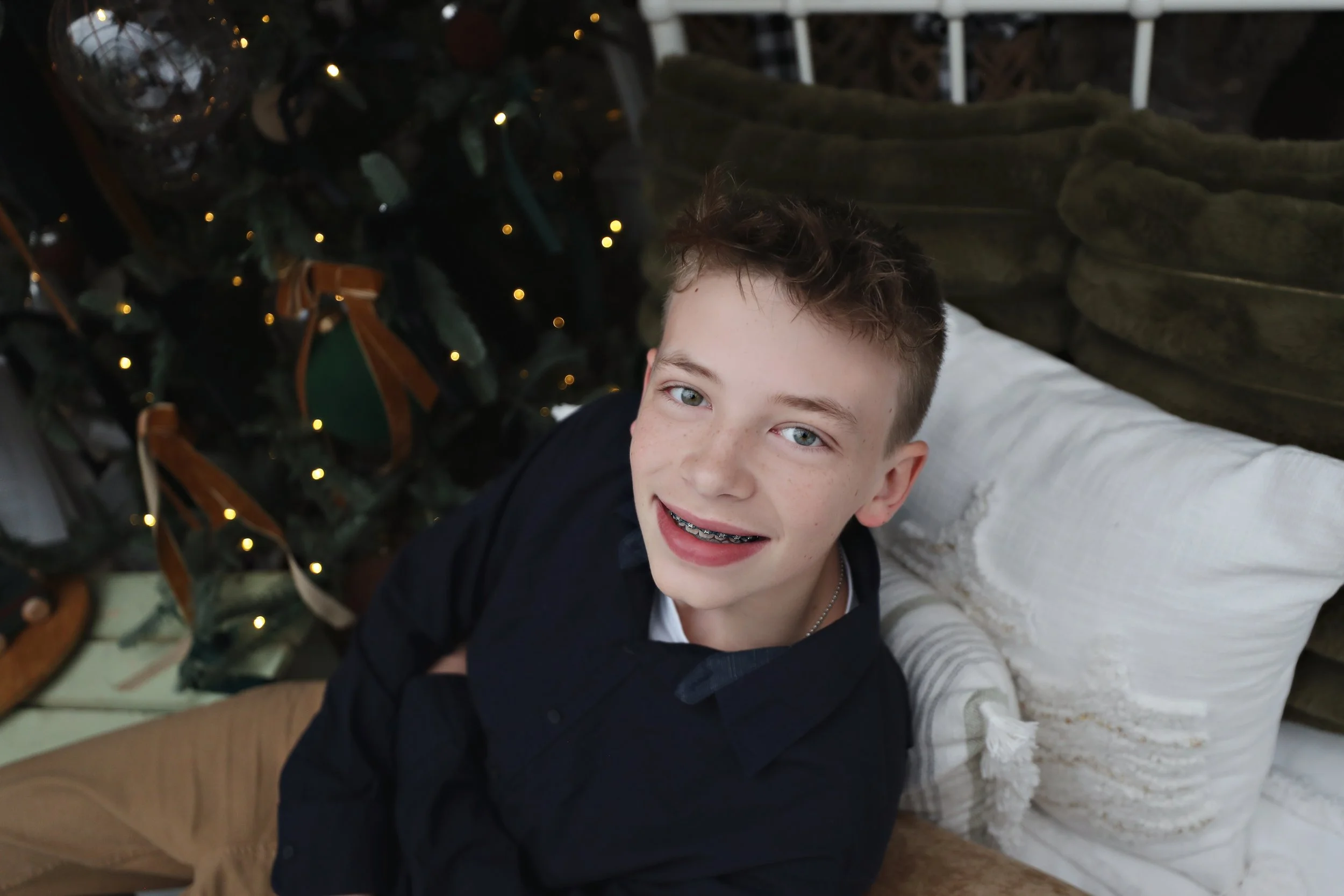 A smiling young boy with braces, sitting on a sofa near a decorated Christmas tree with lights.