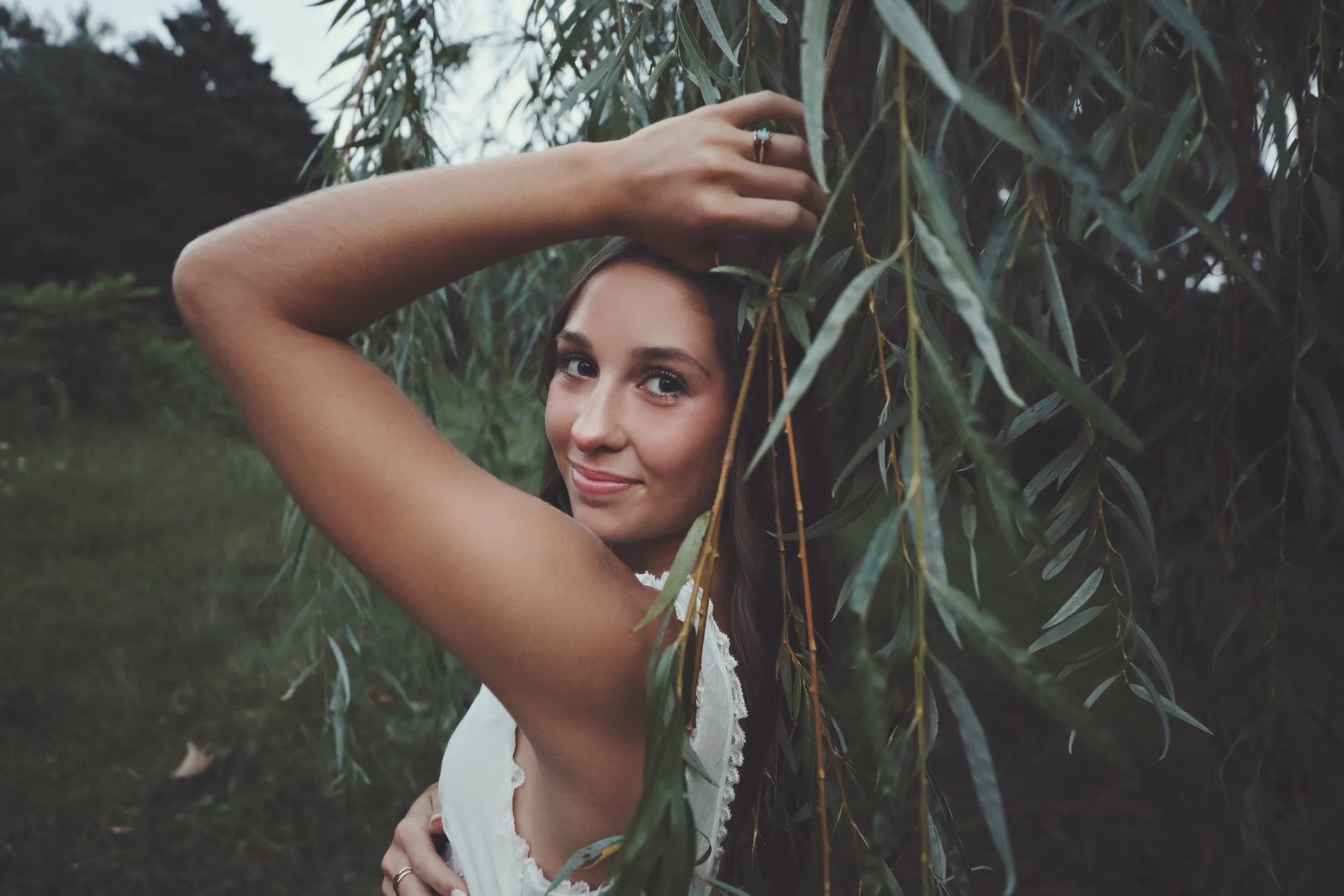 A young woman with light skin and dark hair posing outdoors, touching the leaves of a tree or shrub, with a playful smile.