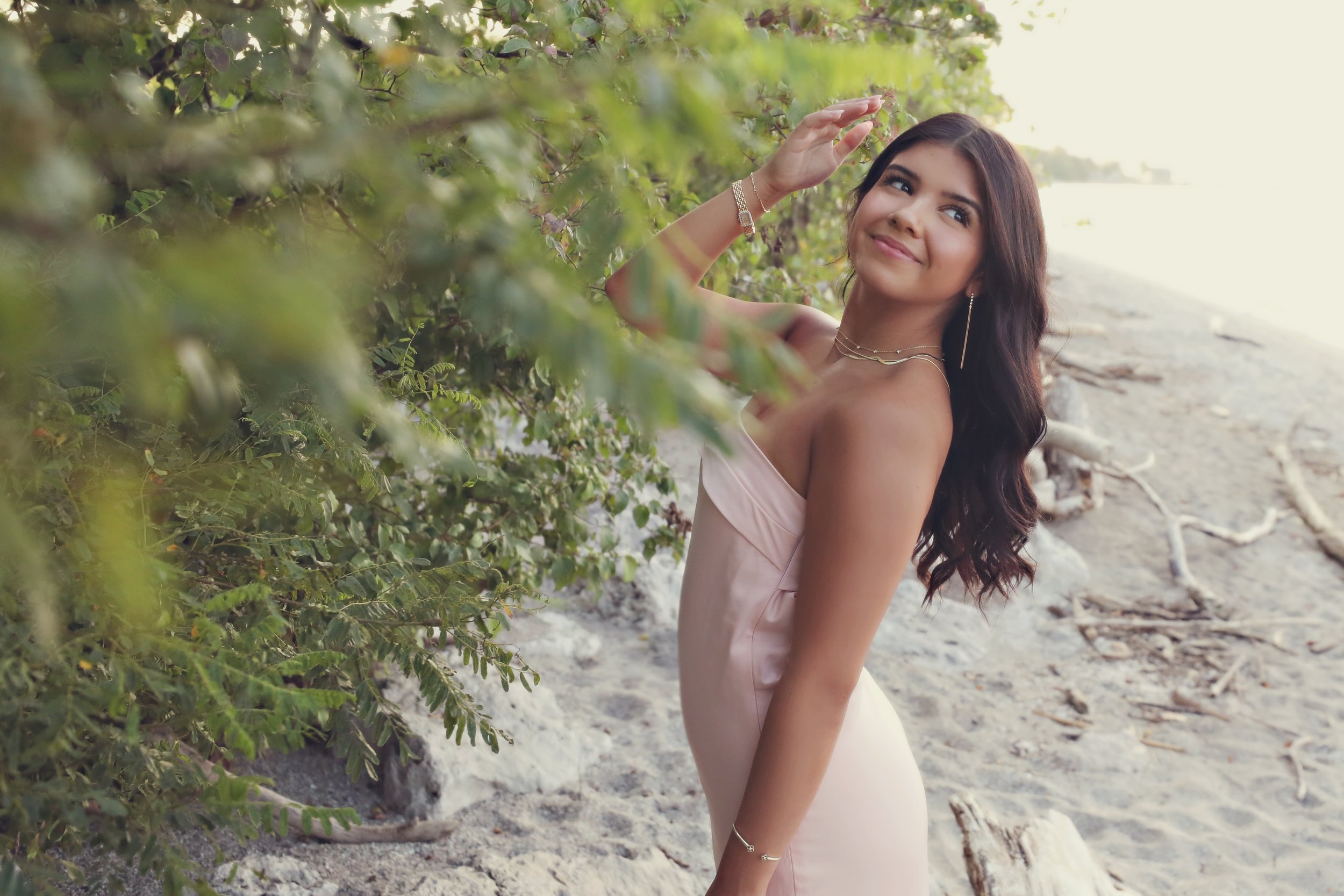 Young woman smiling on a beach, standing near green foliage with sand and driftwood in the background.