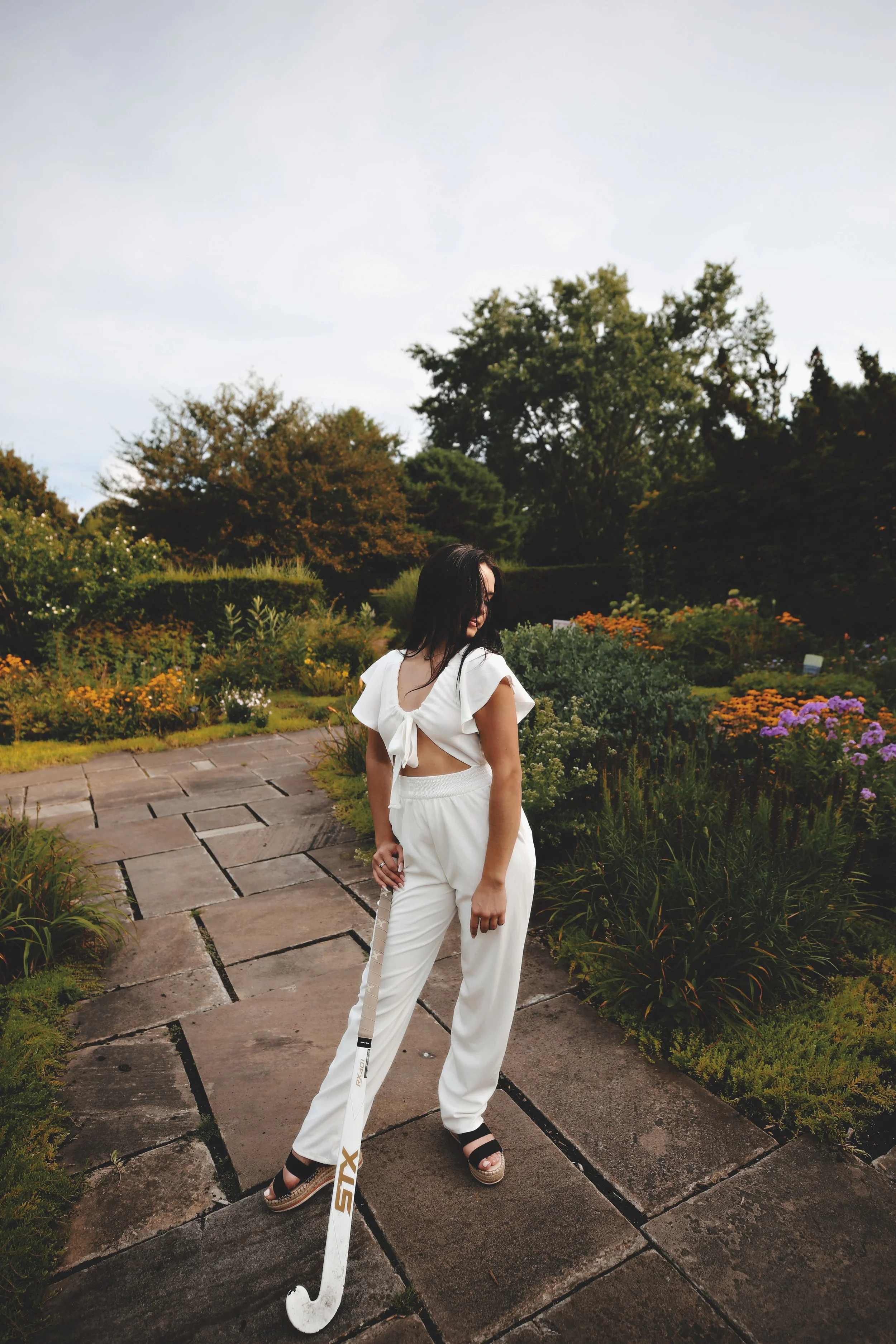 A woman in a white crop top and pants holding a golf club standing on a stone path in a garden with flowers and trees.