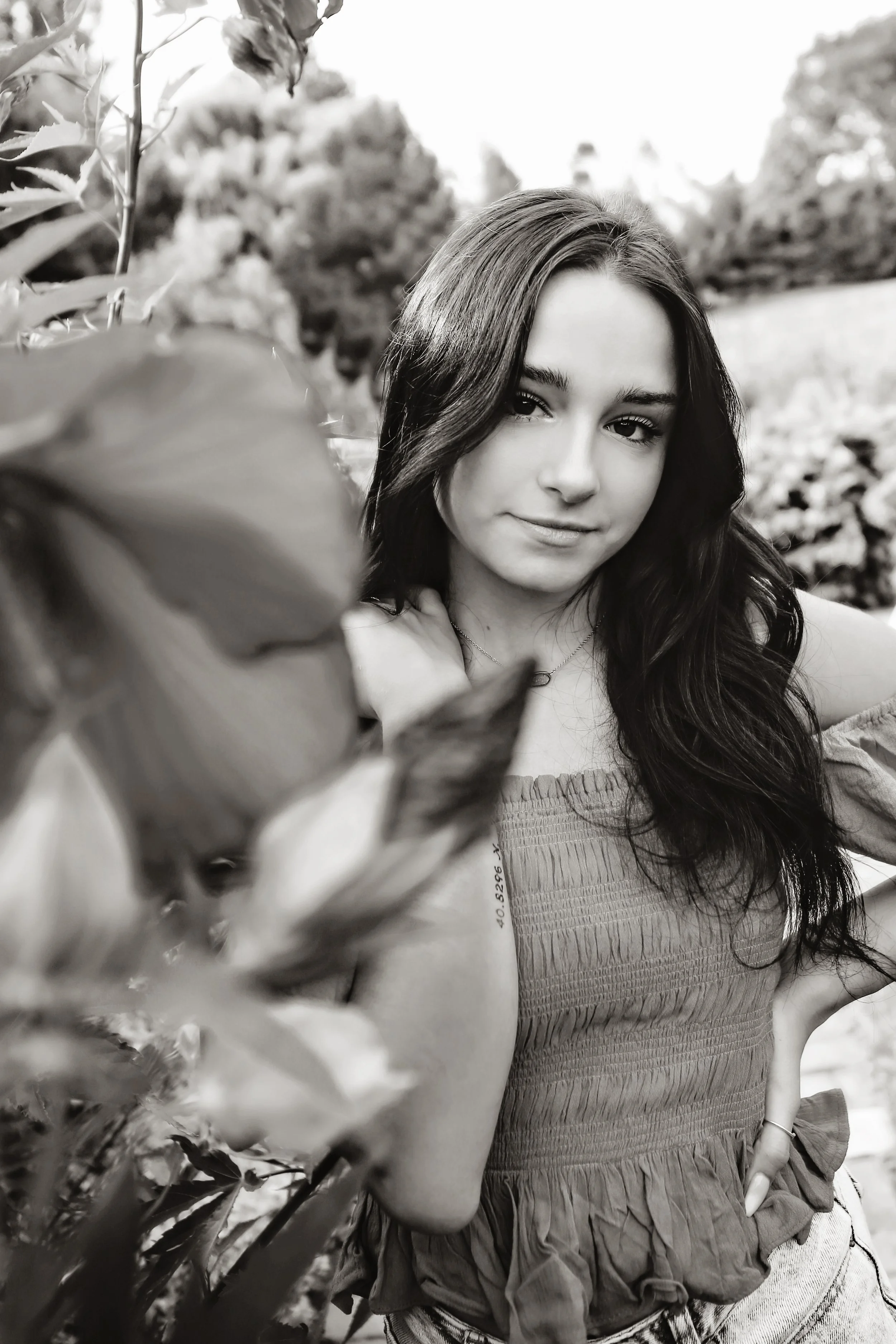 Black and white photo of a young woman with long dark hair, standing outdoors among plants, with trees and sky in the background.