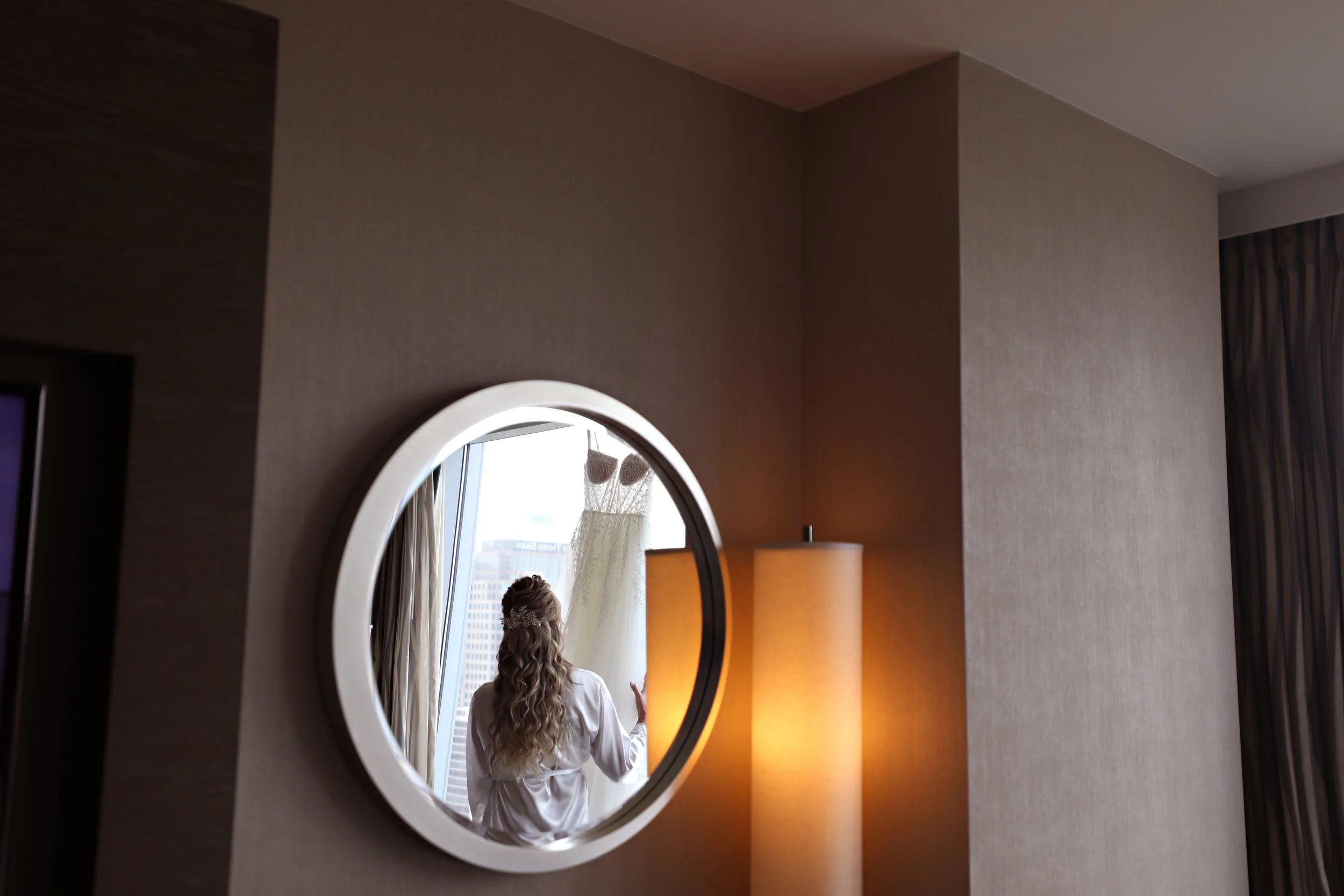 A woman with long, curly hair is looking out a window, her reflection visible in a round mirror on the wall. A white wedding dress is hanging on the window. The room has beige walls and warm lighting from a tall lamp.