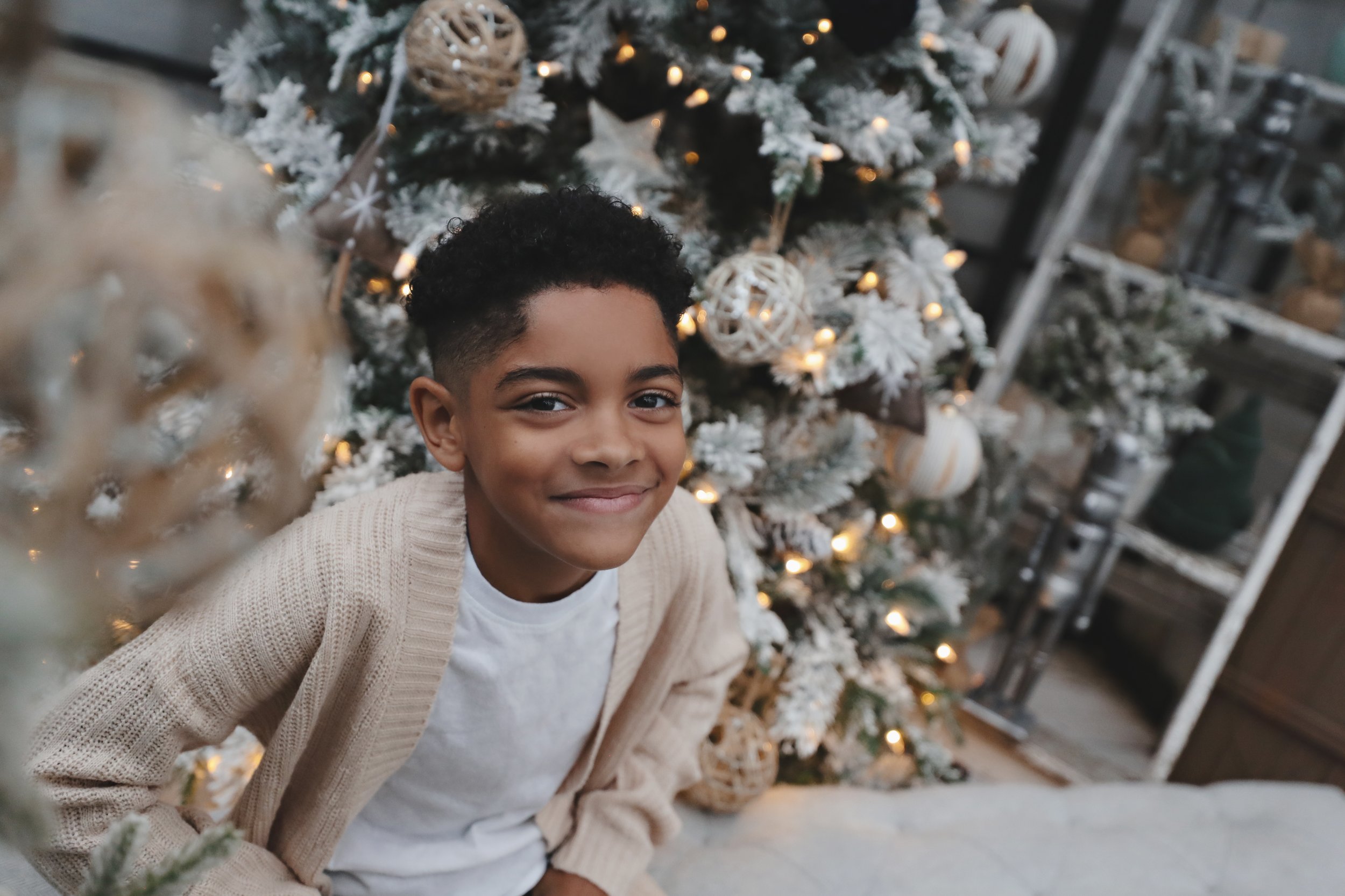 A young boy with short curly hair and a beige cardigan smiling near a decorated Christmas tree.