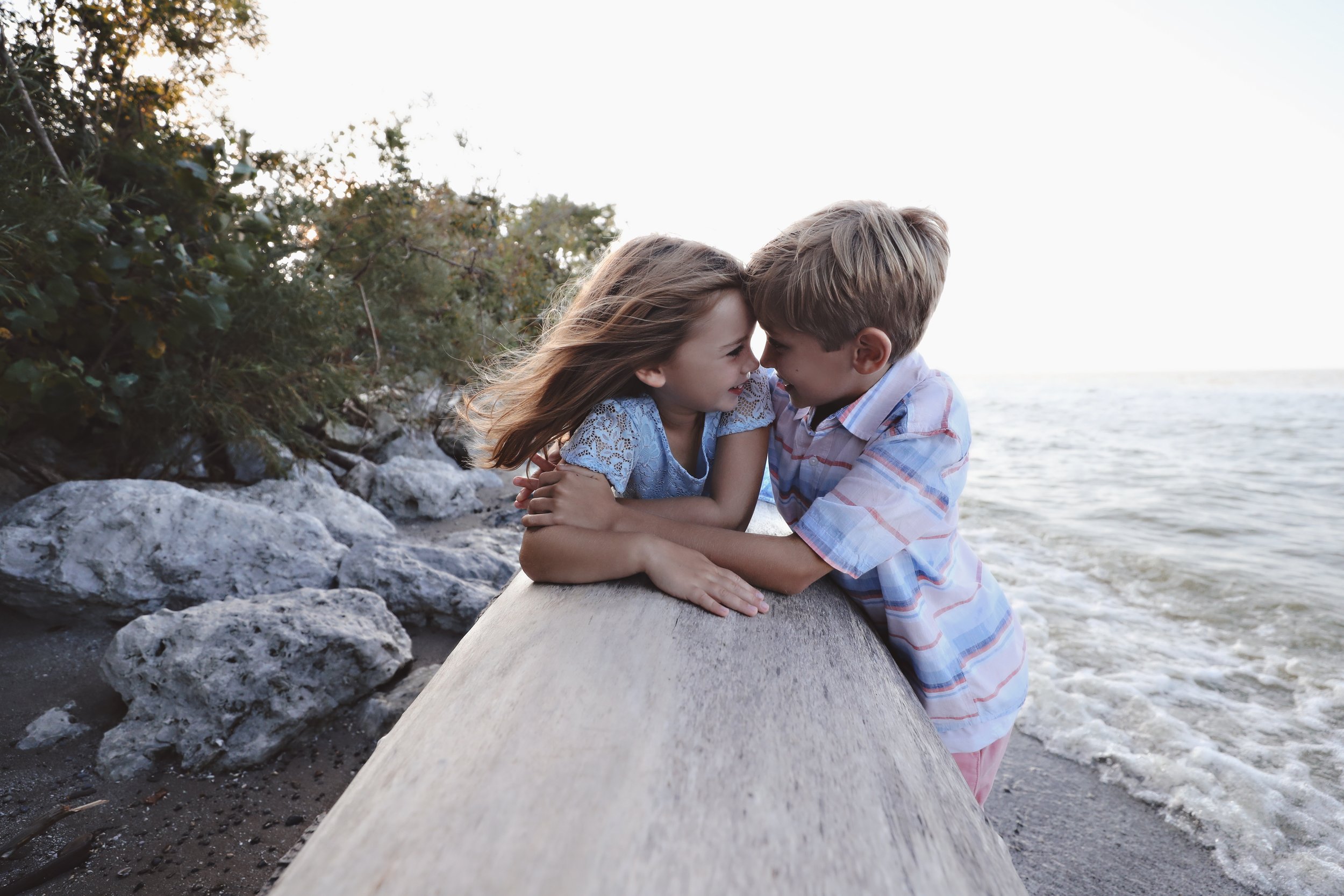 A young boy and girl near the shoreline, leaning on a large log, touching foreheads, with water and rocks in the background.