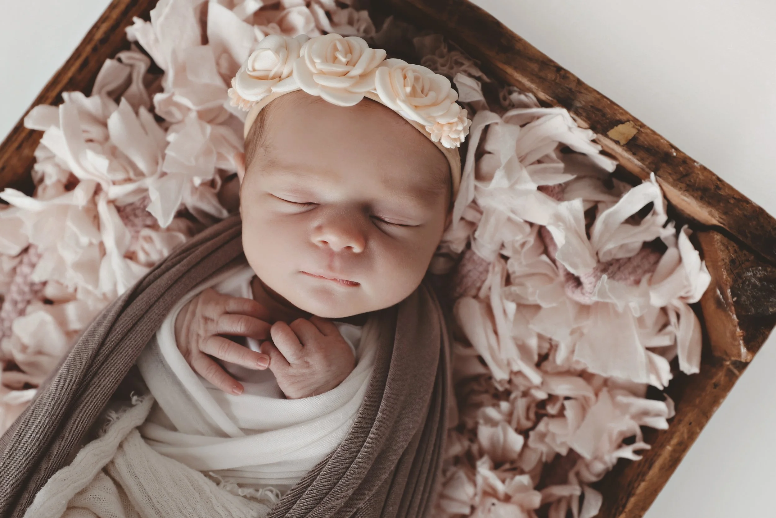 A sleeping baby with a floral headband, lying on pink petals in a wooden basket.