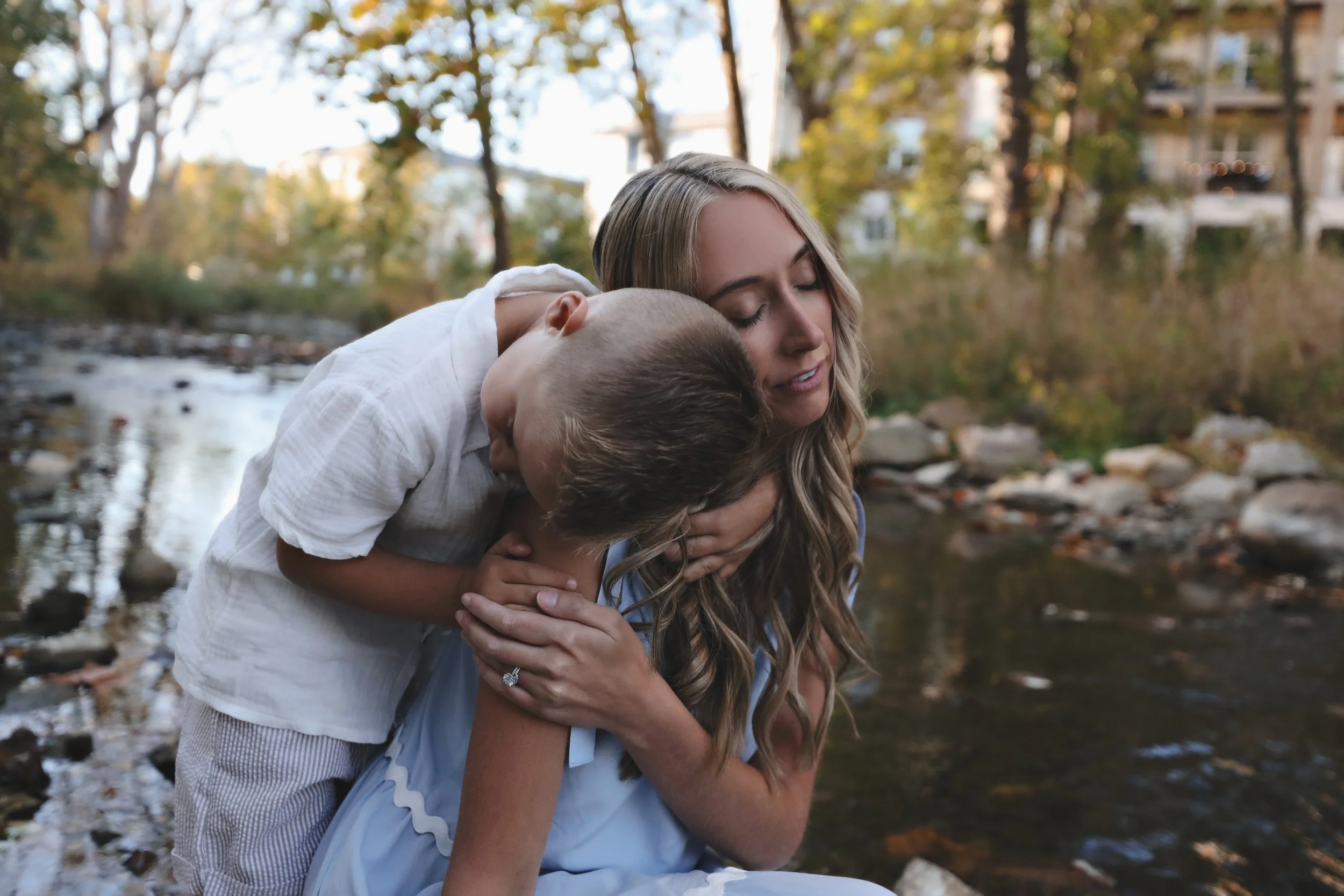 A woman and two children by a creek in fall; one child is on her back, hugging her, the woman has long blonde hair and closed eyes, feeling affection in a peaceful outdoor setting.
