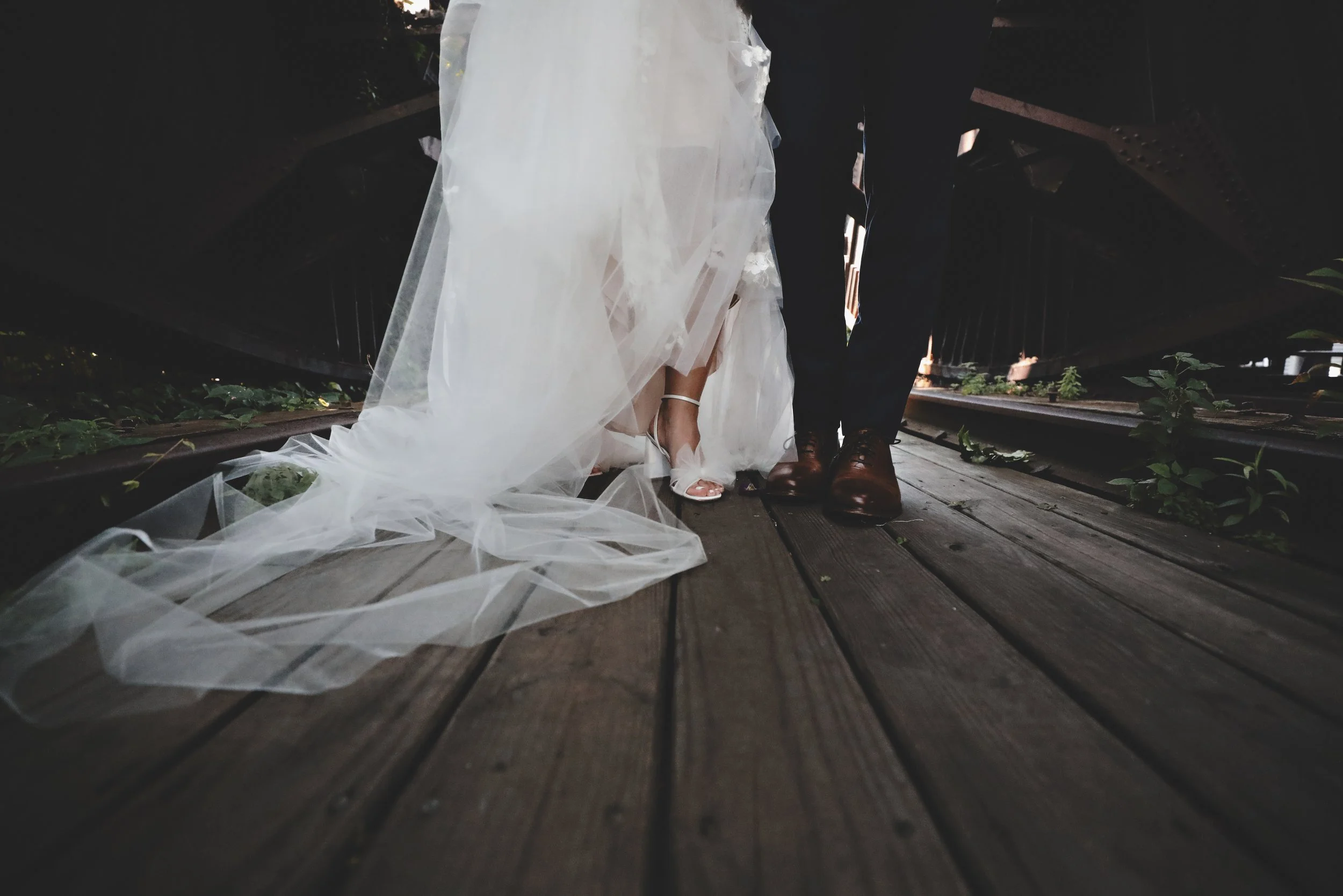 Bride and groom standing on a wooden train track, seen from a low angle.