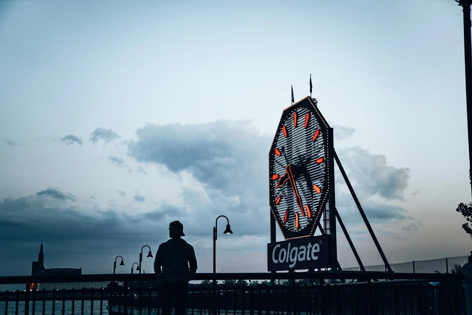 Silhouette of a person standing by a water body at dusk with a large illuminated clock tower showing the time as approximately 8:28, and a "Colgate" advertisement below.