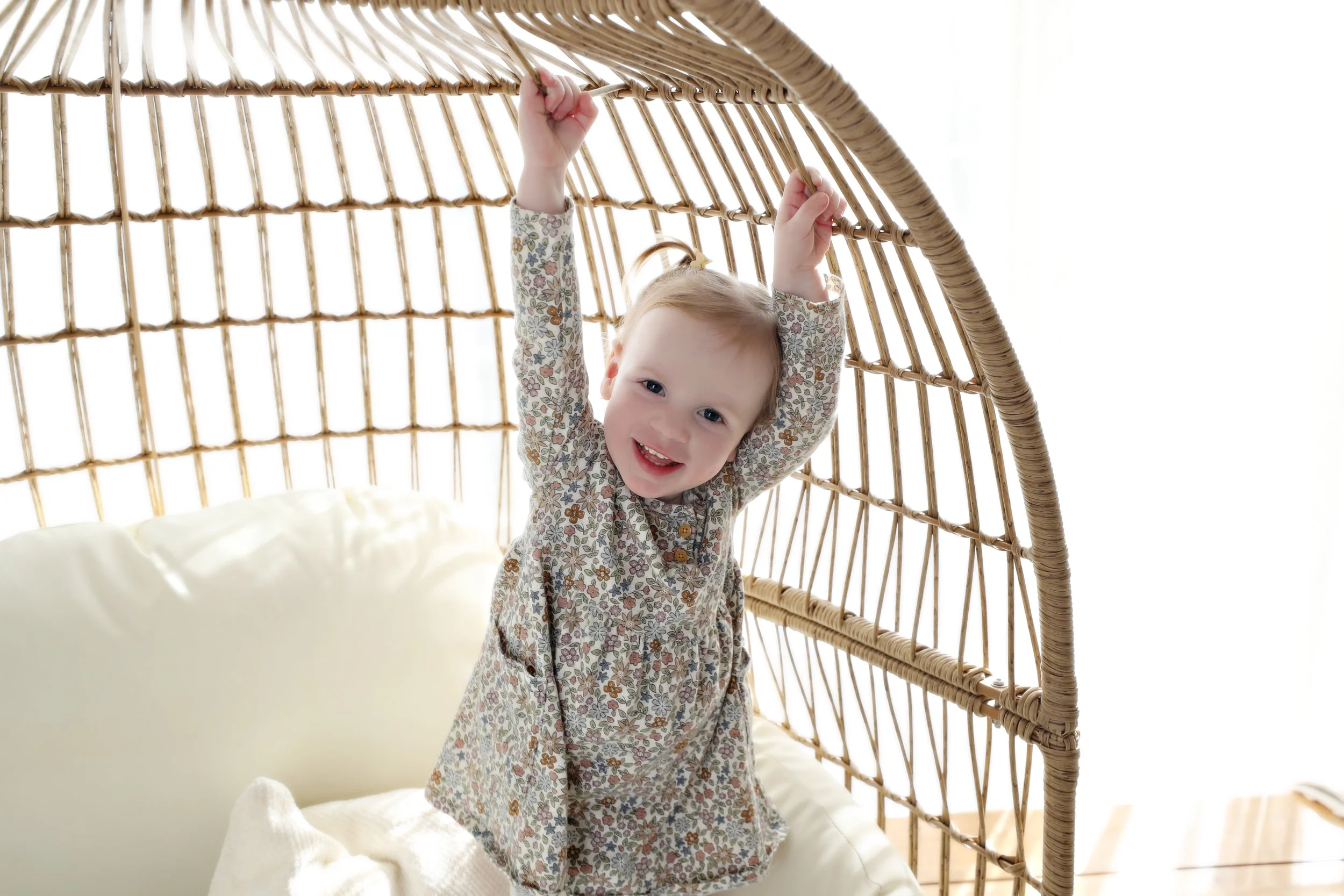 A smiling young girl with blonde hair and blue eyes, wearing a floral dress, hanging and playing inside a wicker hanging chair.