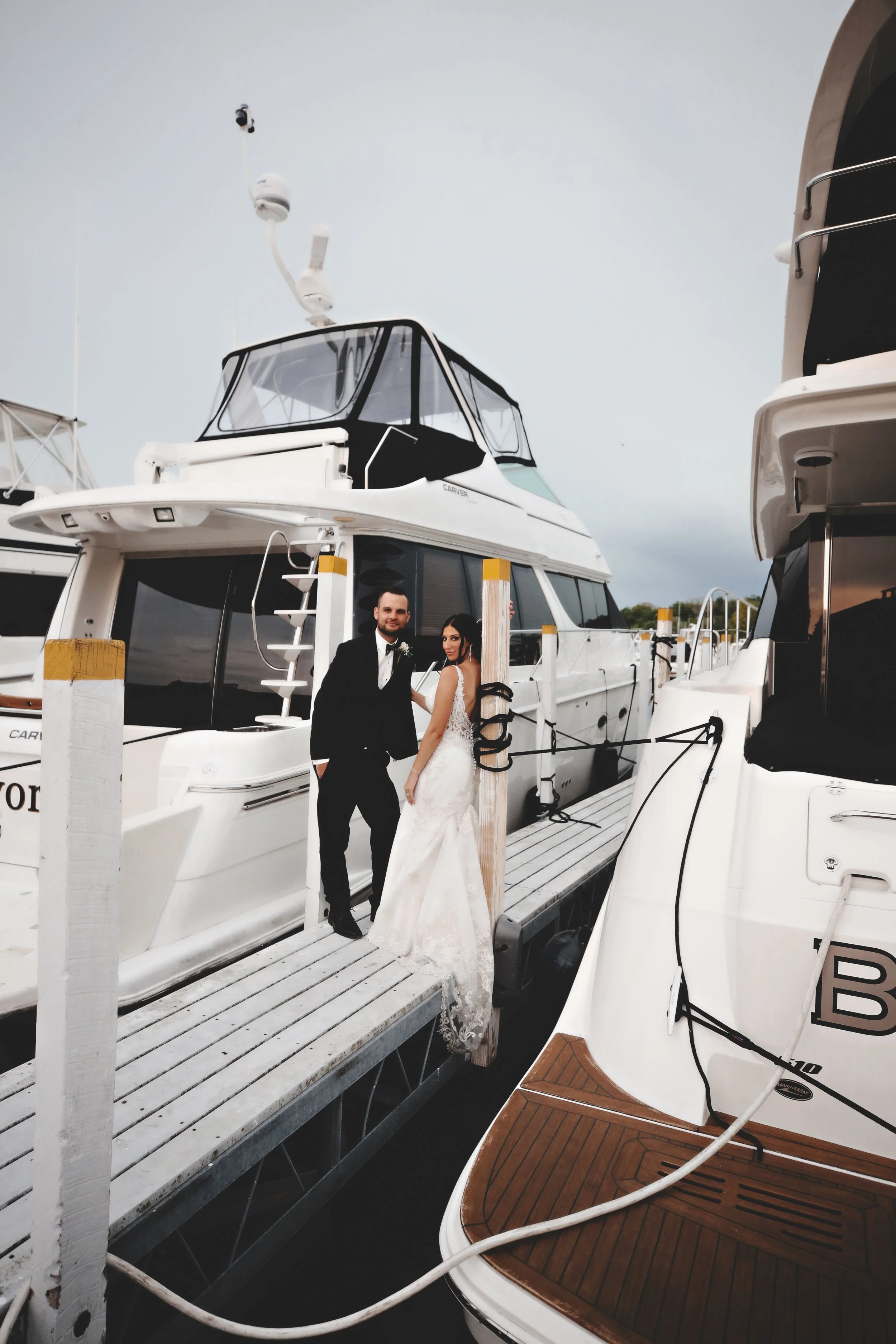 A bride and groom standing on a dock between yachts, with the groom in a black tuxedo and the bride in a white lace wedding gown, by a boat.