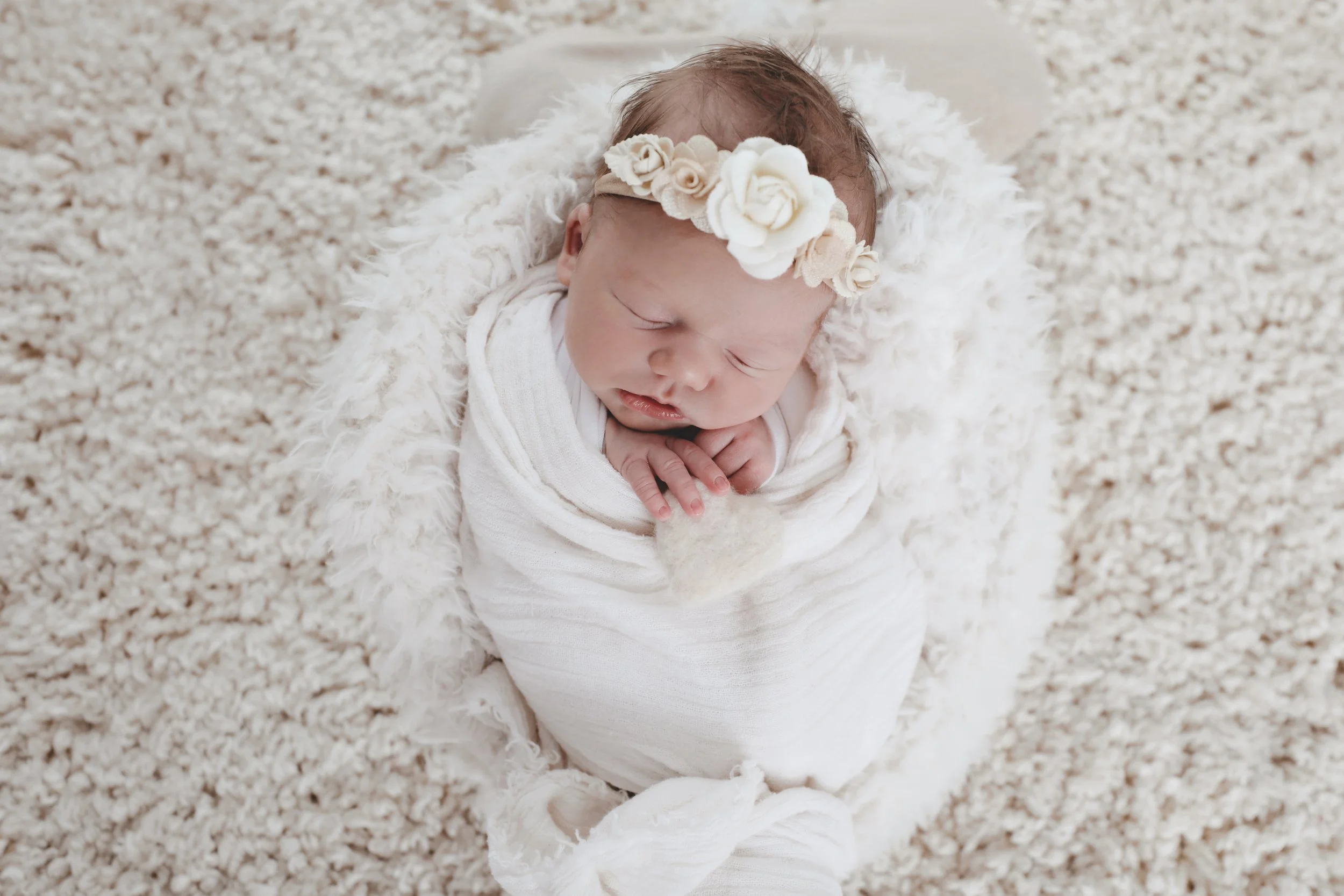 A sleeping newborn baby wrapped in a white blanket, wearing a floral headband, lying on a plush, cream-colored surface.