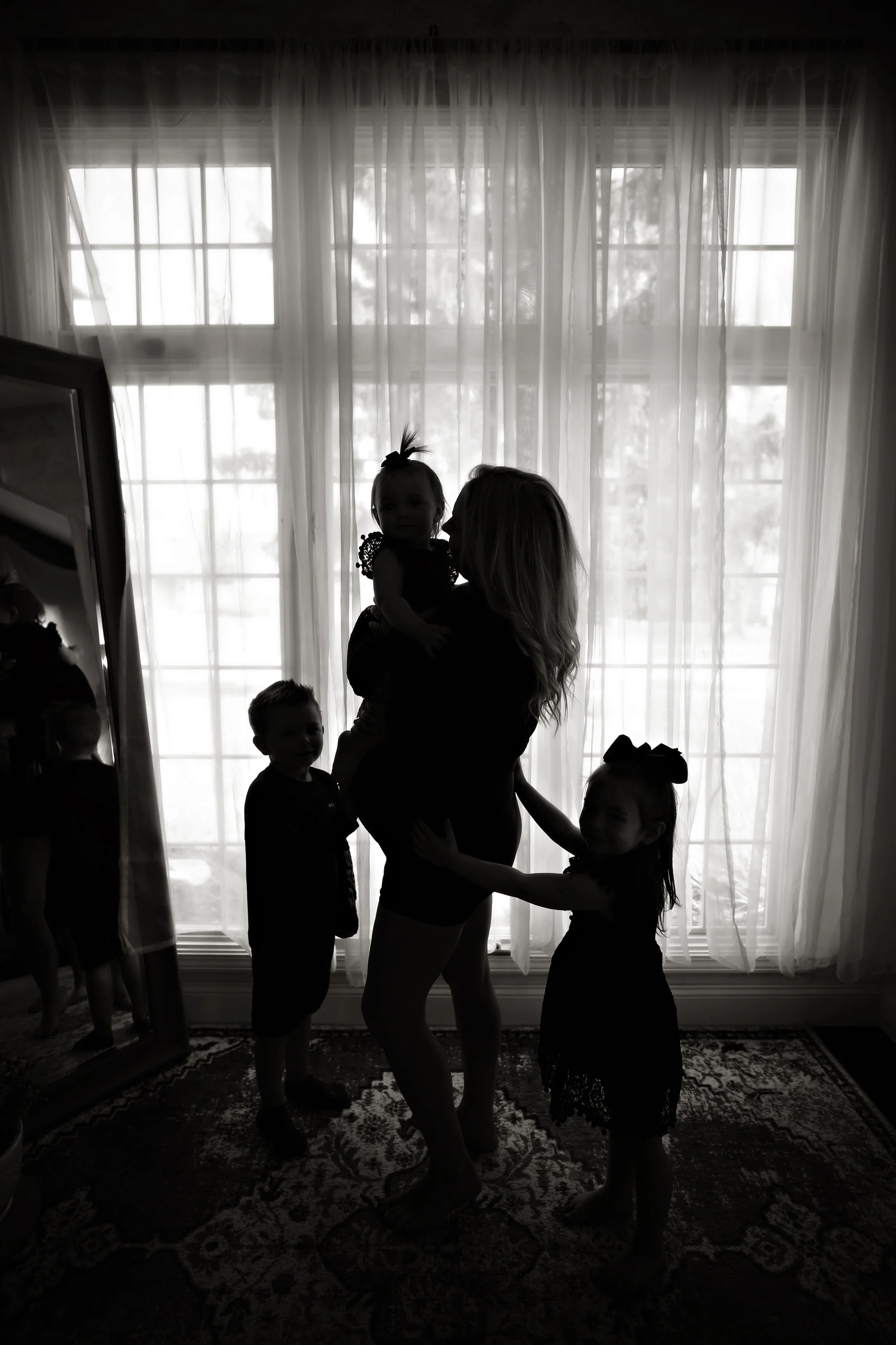 Silhouette of a woman holding a child while standing on an ornate rug in front of large windows with sheer curtains. Three other children stand nearby, with one reflected in a large mirror on the left.