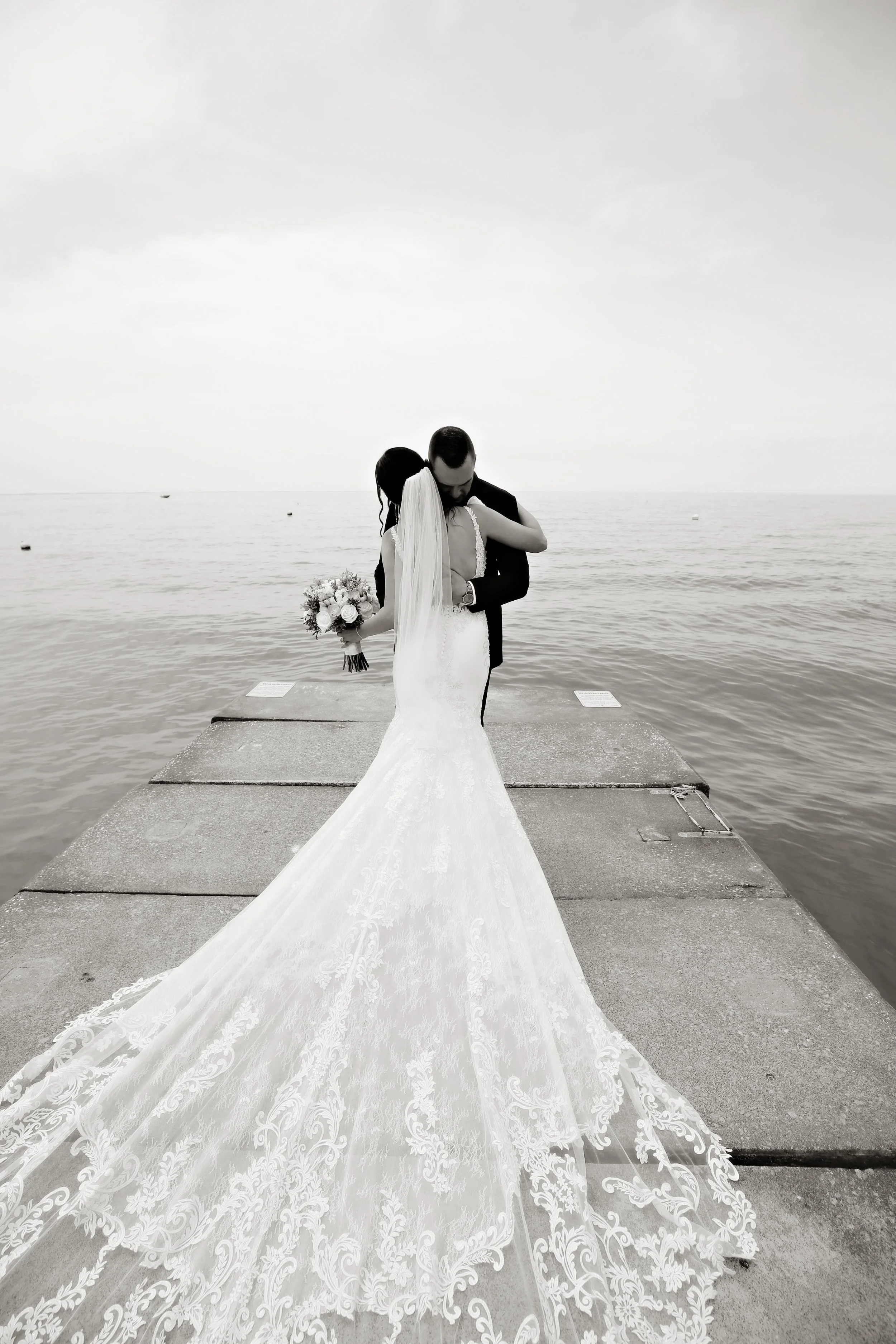 A bride and groom embracing on a dock overlooking the water, with the bride holding a bouquet, in black and white.