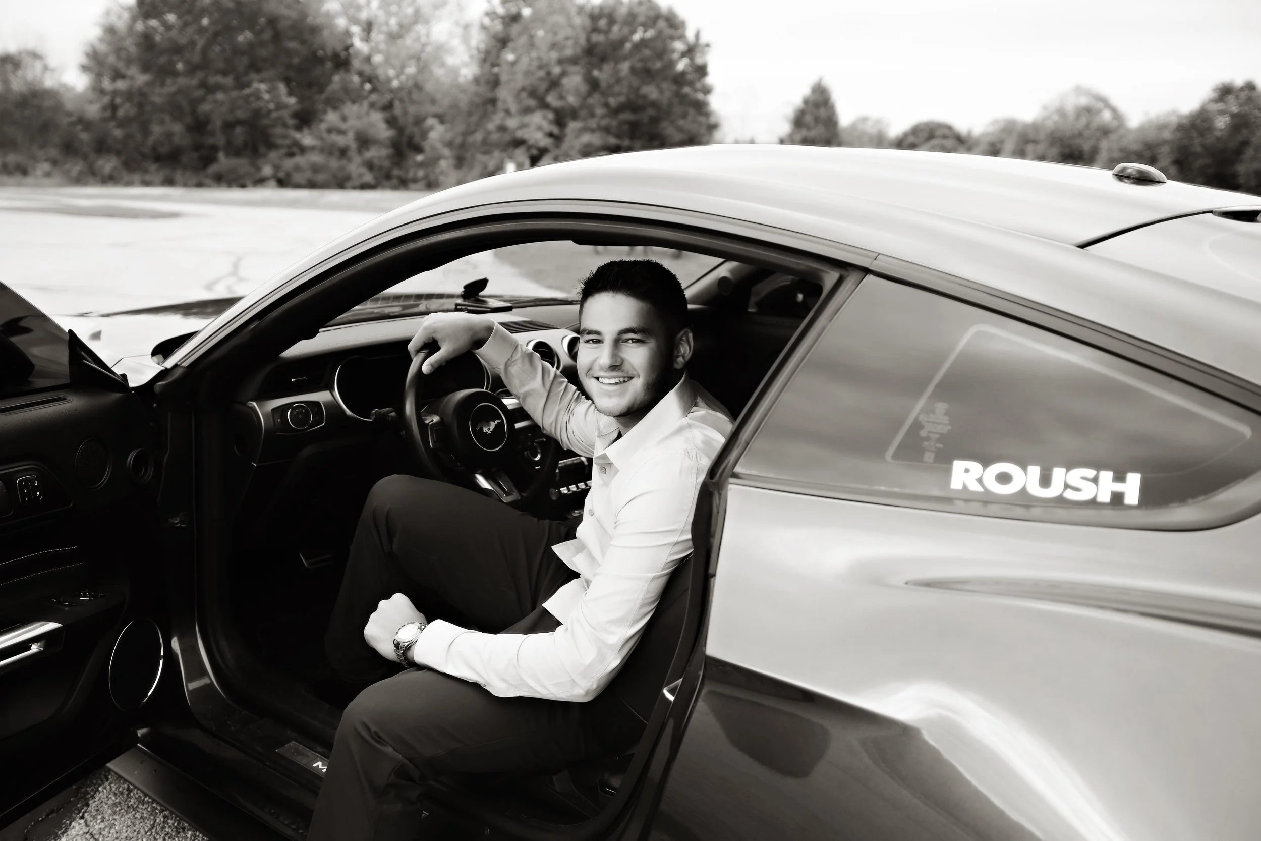 A smiling man in dress shirt and watch sitting in the driver's seat of a sports car, with the door open, in an outdoor setting. The car is a Roush model, with a logo on the side window. The background shows trees and an open sky.