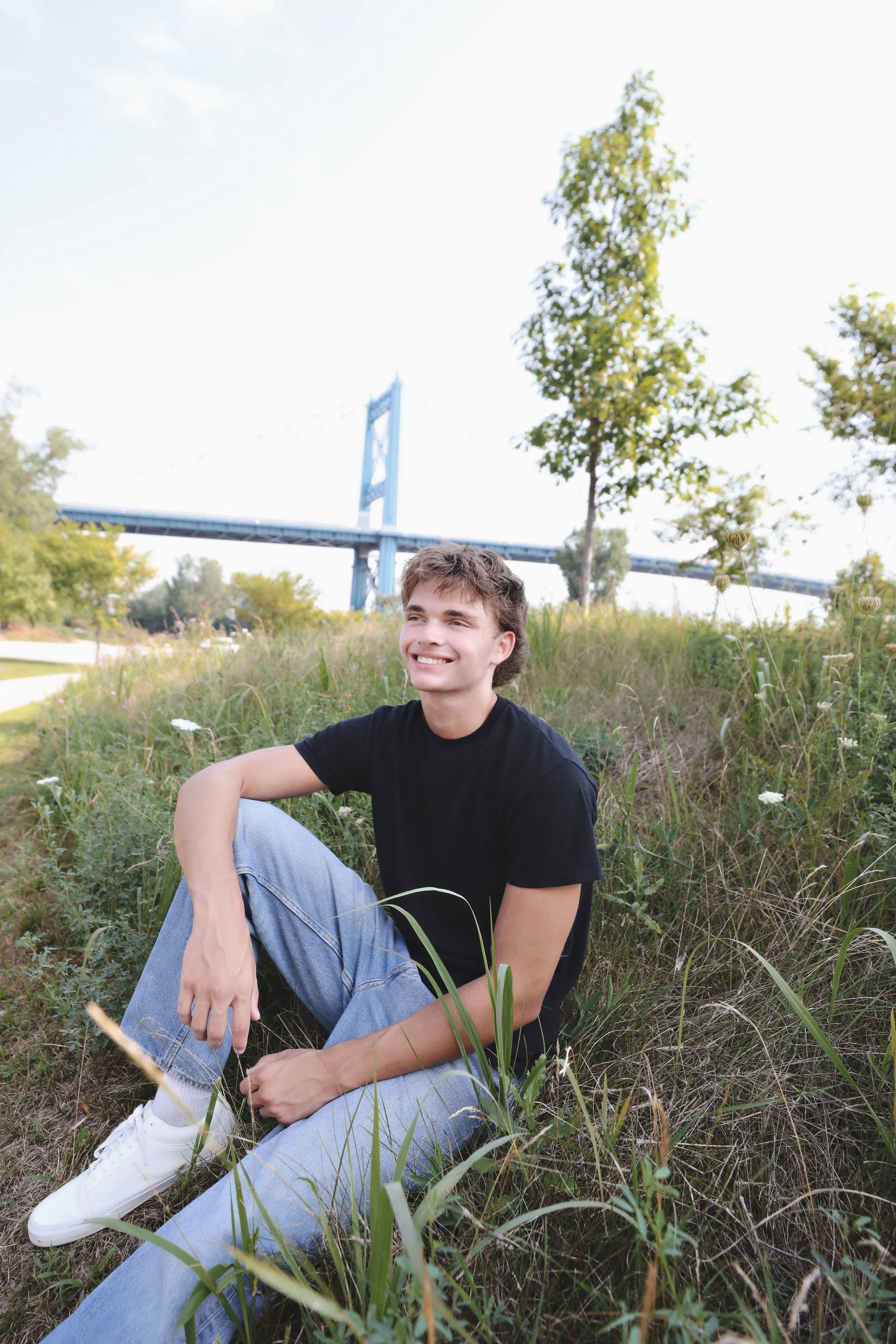 A young man sitting outdoors on a grassy area, smiling, with a bridge and trees in the background.