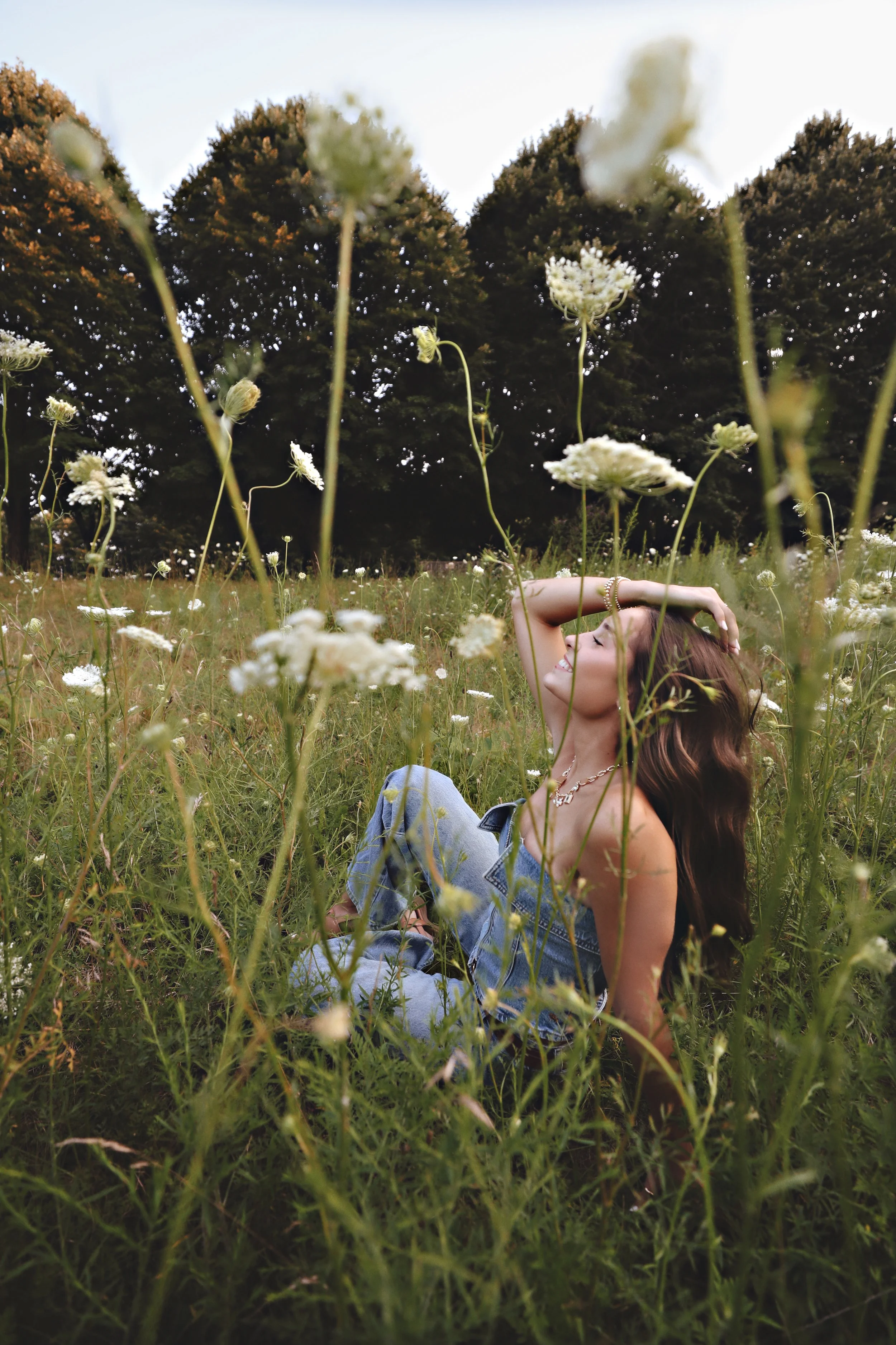 A woman sitting in a field of wildflowers, smiling with her eyes closed, with trees in the background.