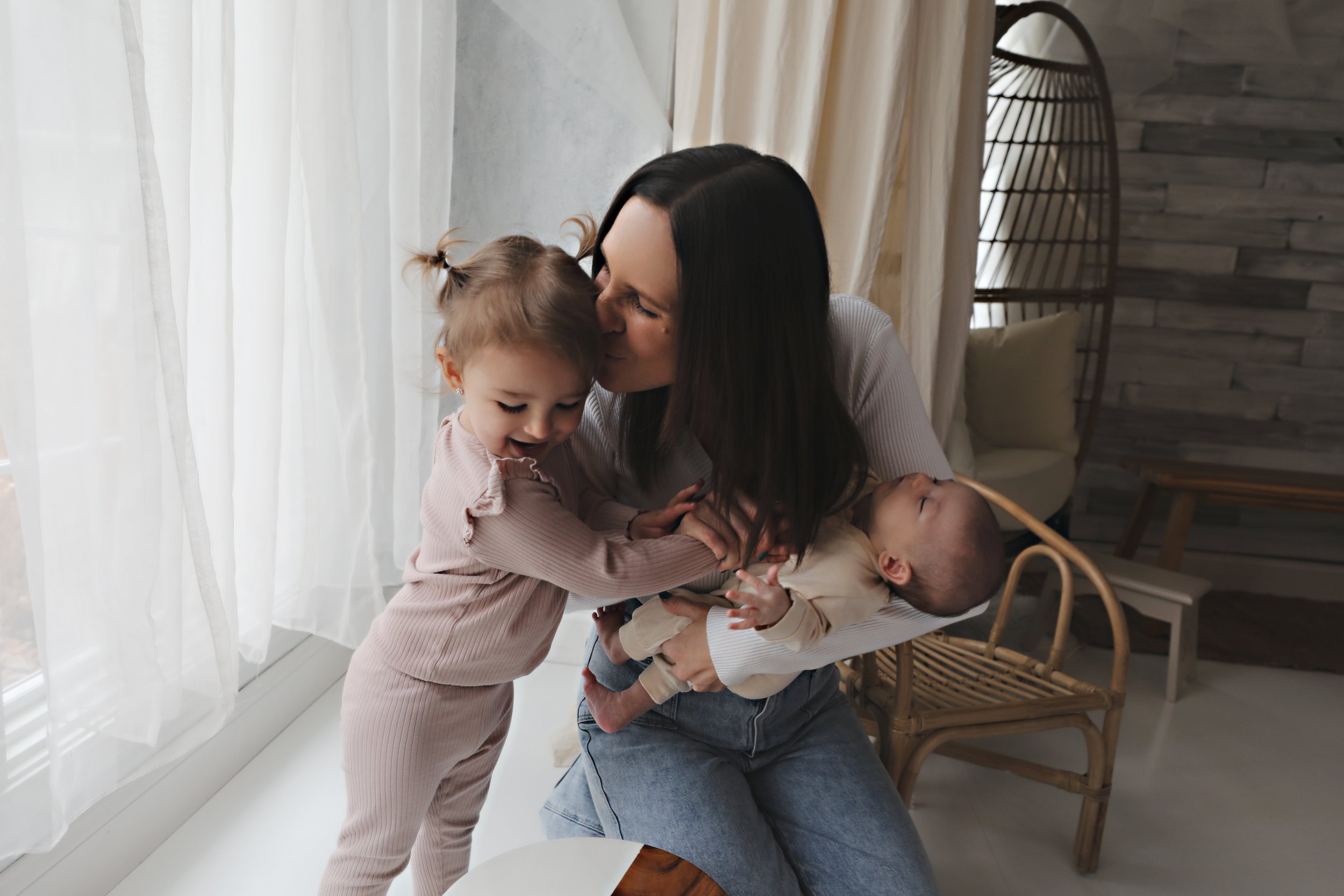 A woman with long dark hair holding a baby and kissing a young girl on the cheek in a sunlit living room.