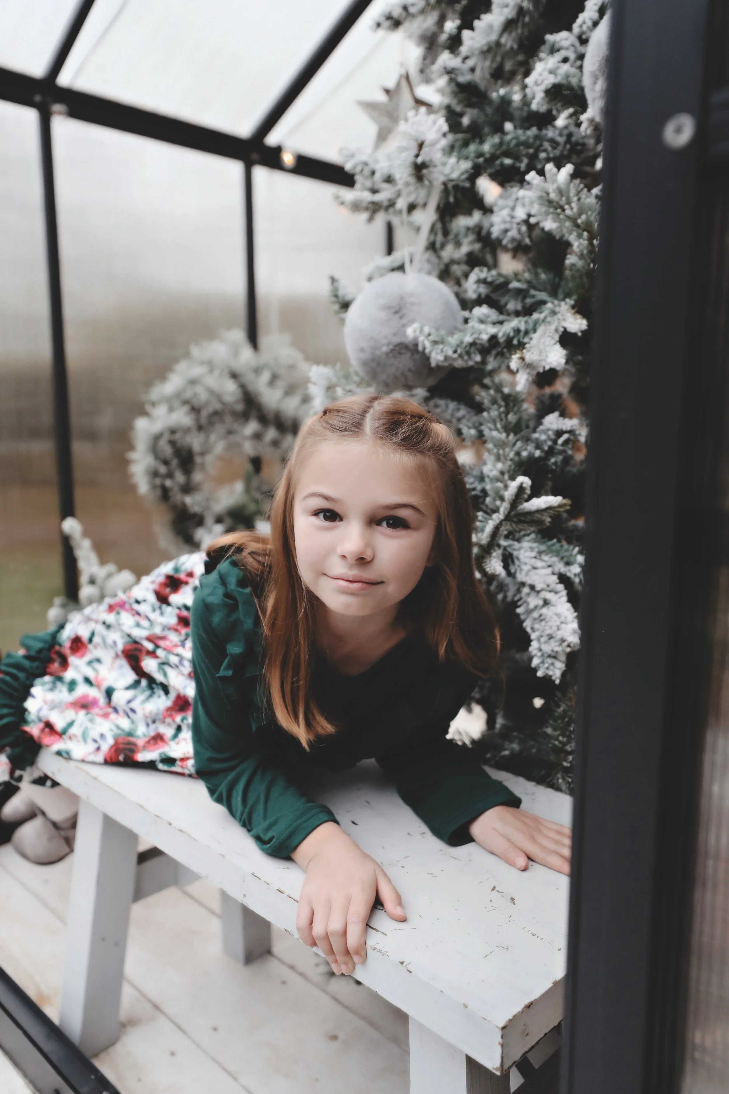 A young girl with light brown hair lying on a white wooden bench inside a greenhouse, next to a snow-covered Christmas tree decorated with a star and ornaments.