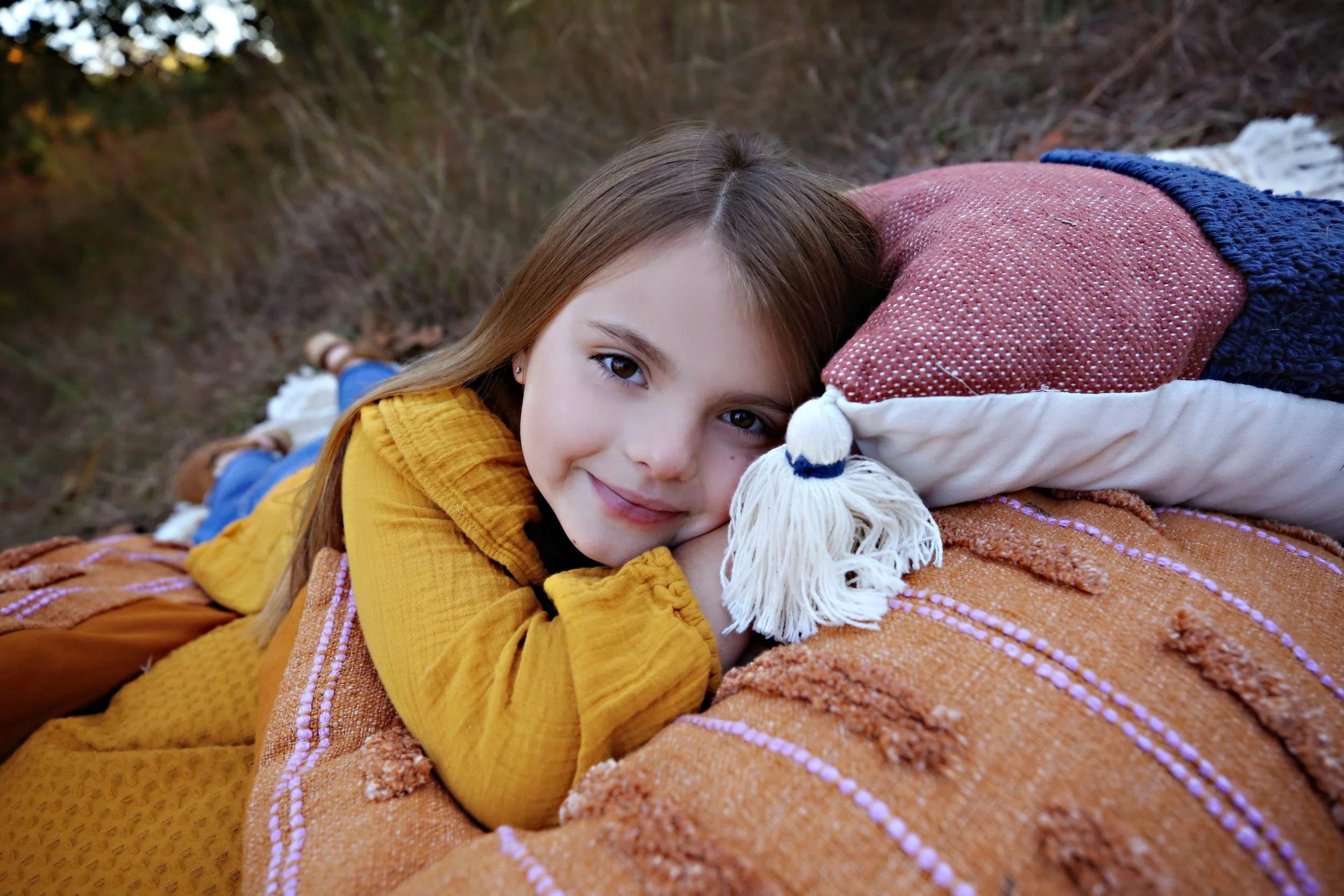 A young girl with long brown hair, lying on her side on a bed of blankets outdoors, resting her head on a pillow, smiling at the camera.