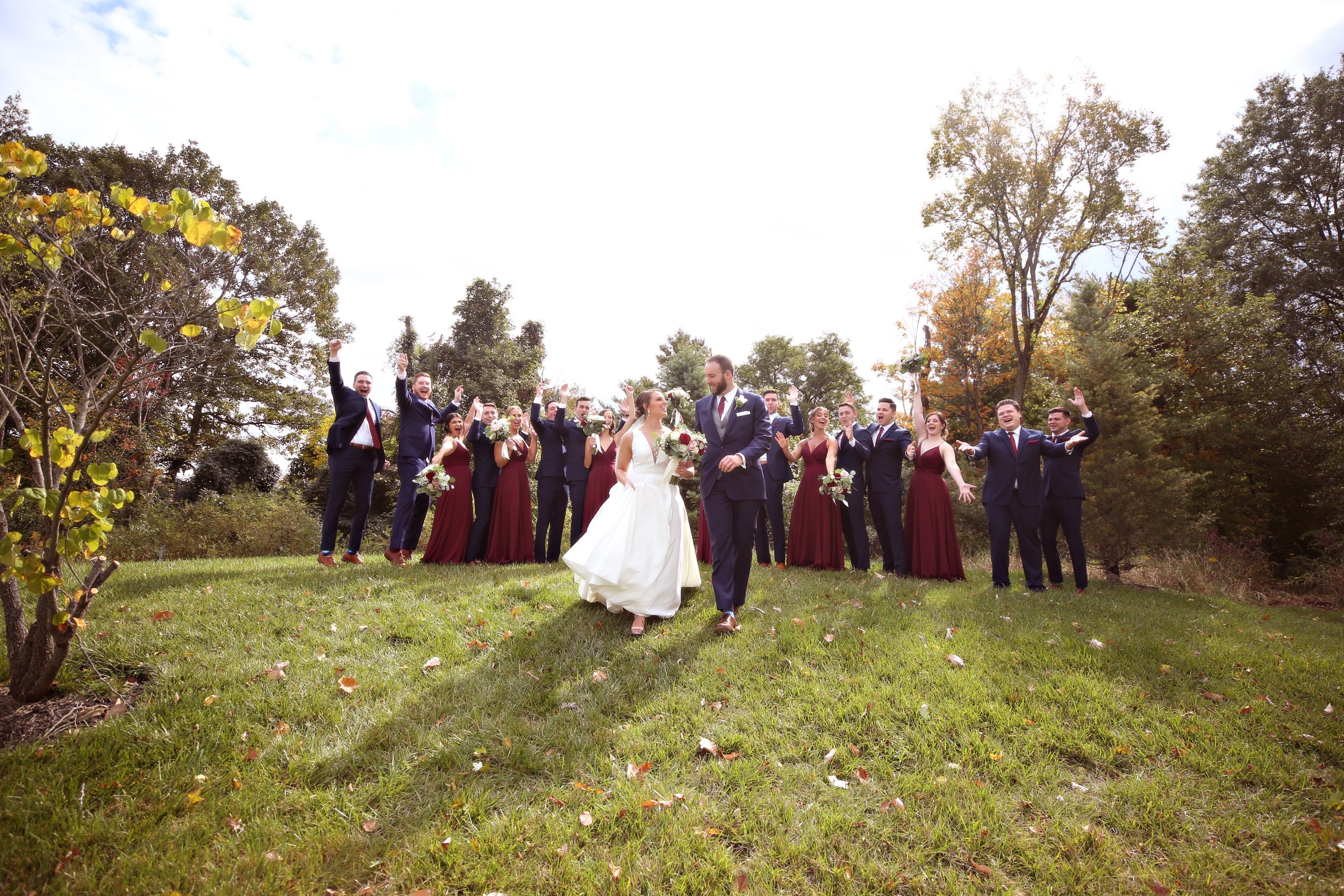 A wedding party outdoors in a grassy area with trees, featuring a bride and groom in the center, surrounded by bridesmaids in burgundy dresses and groomsmen in navy suits, celebrating and smiling.